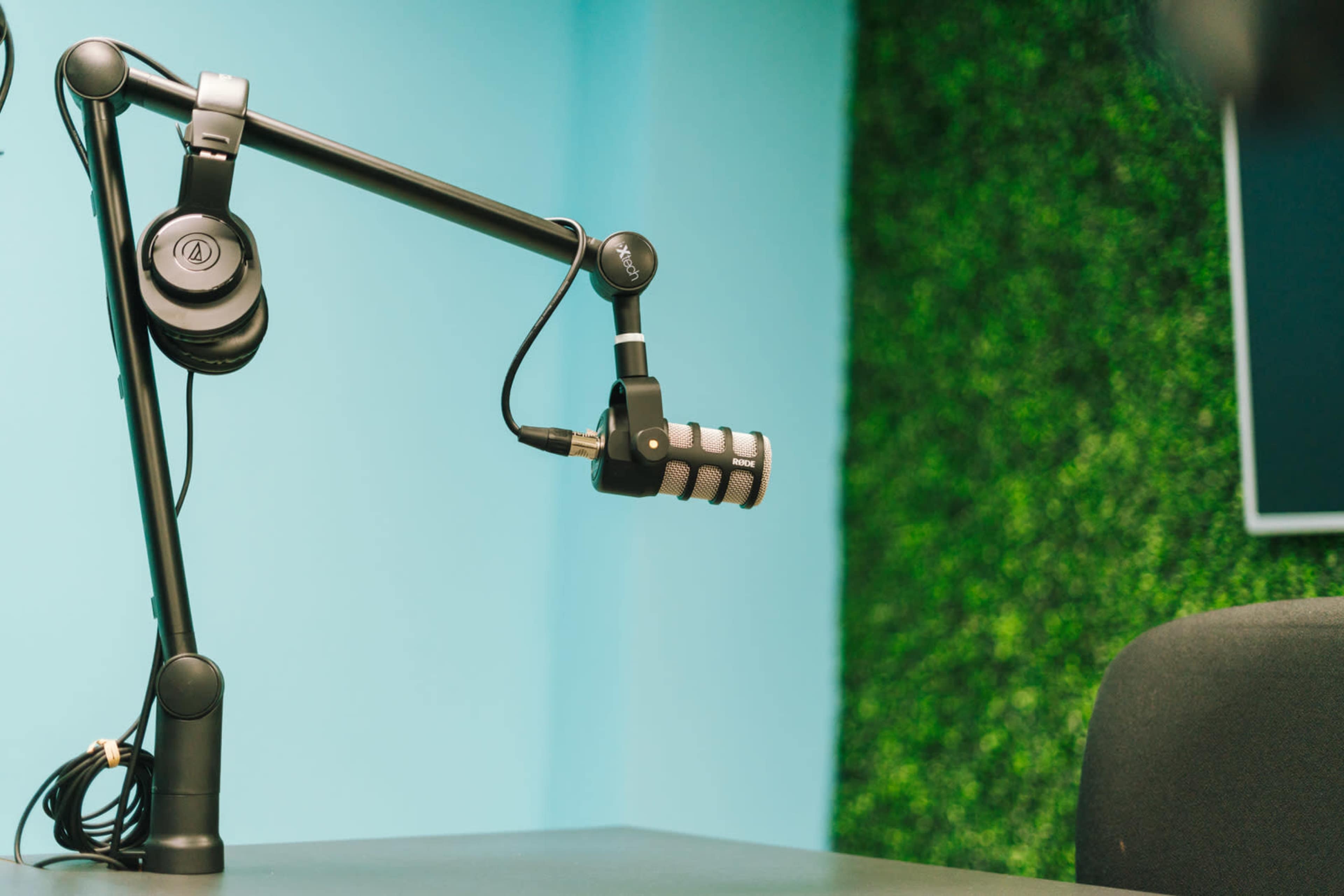 A microphone attached to a black adjustable arm is positioned on a desk in a studio with a blue wall and a green textured background.