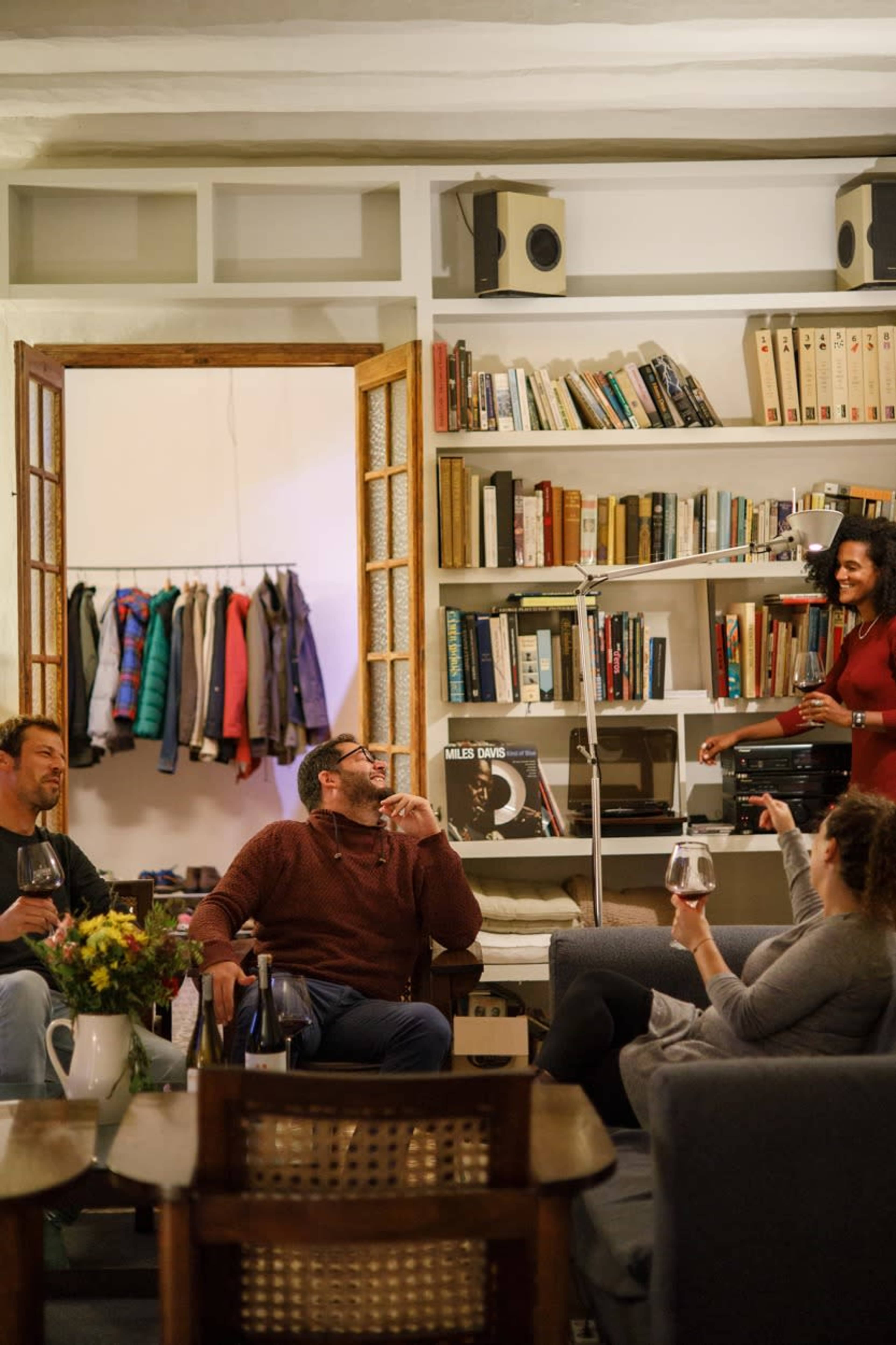 A group of four people socializes in a cozy living room filled with bookshelves, while one person serves drinks.