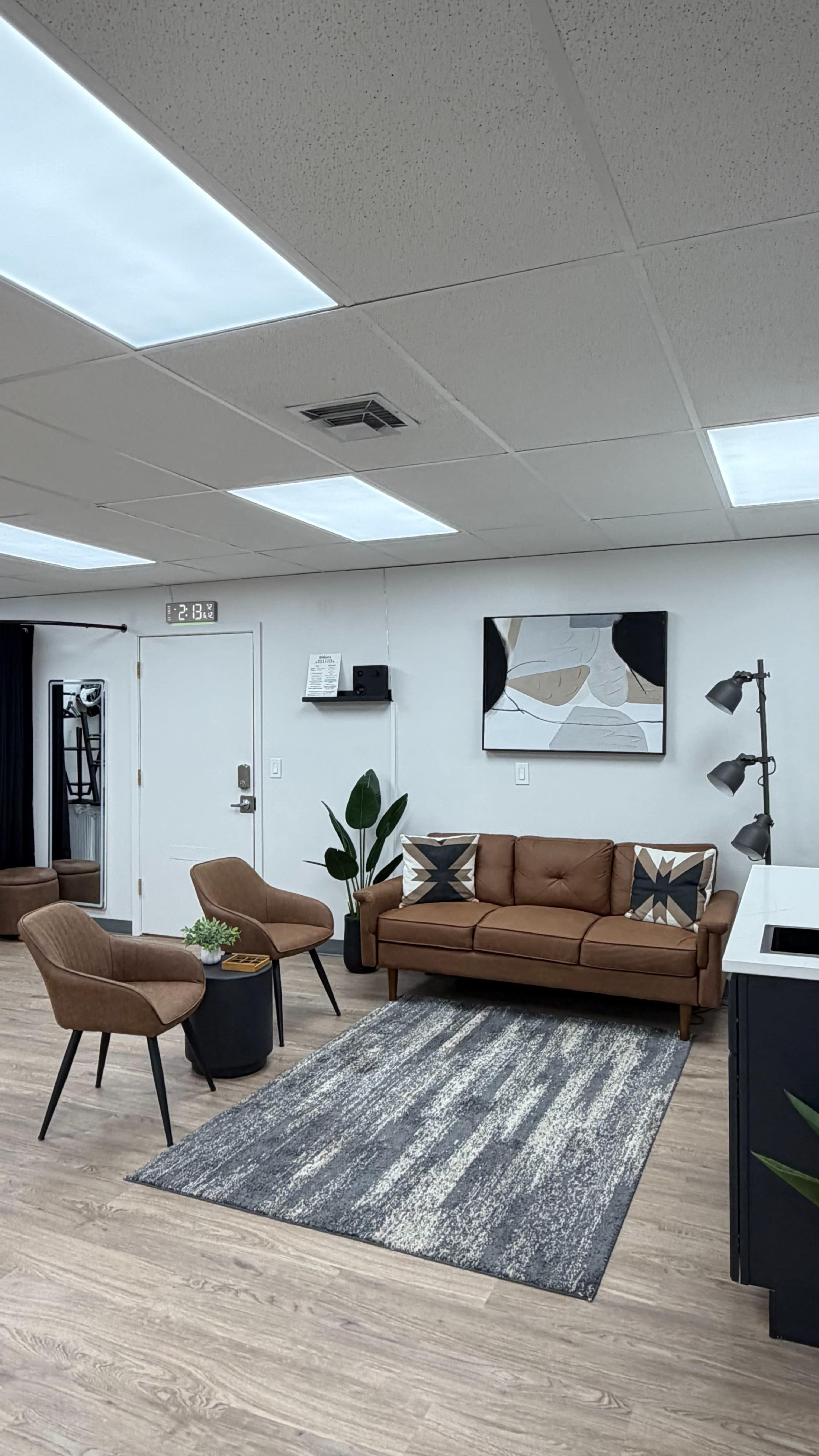 The image shows a modern interior design featuring a brown leather sofa, two armchairs, a black floor lamp, and a decorative area rug, all positioned in a well-lit space.