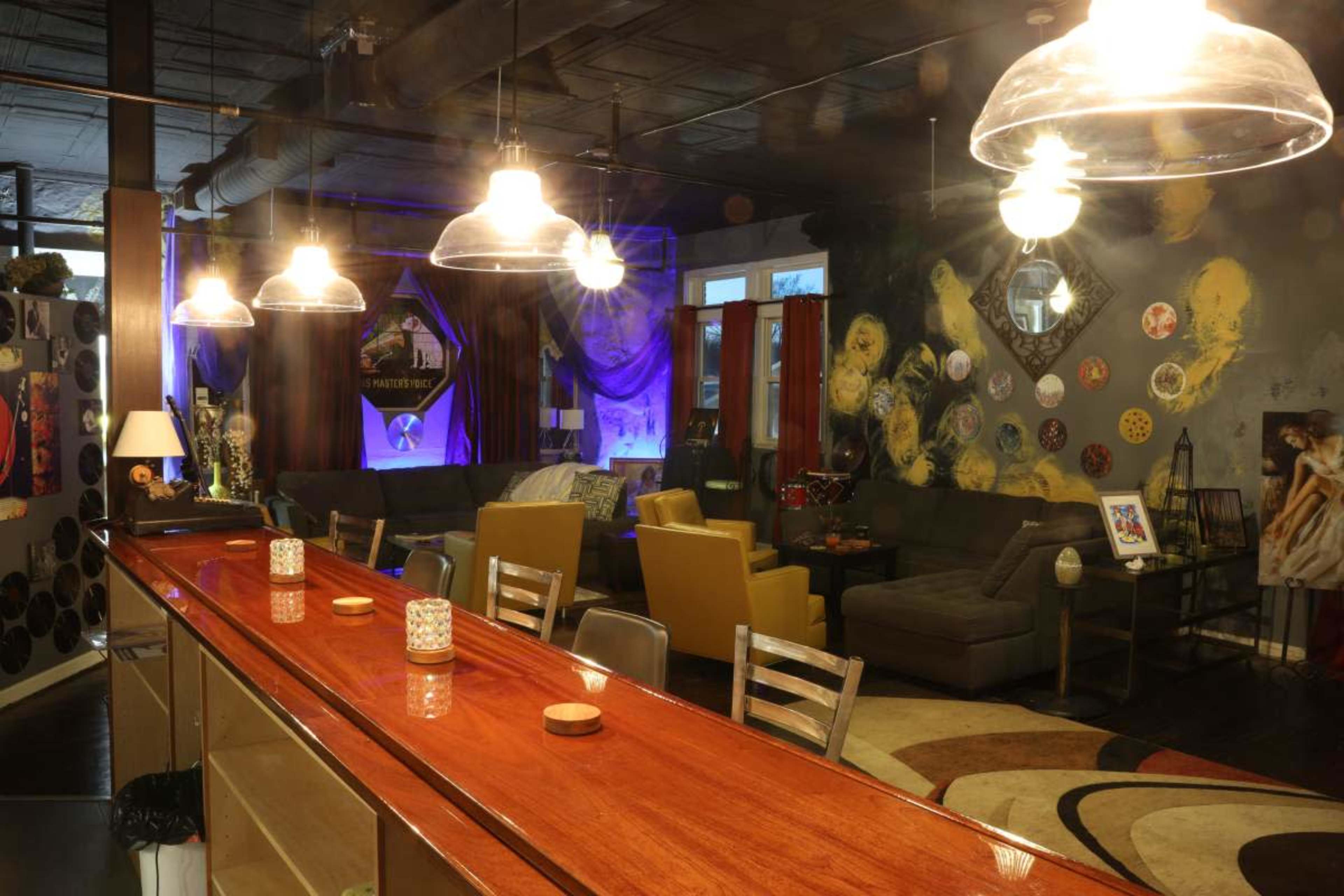 The scene shows a modern lounge area with a long wooden bar, several seating arrangements, and colorful artwork on the walls, illuminated by hanging lights.