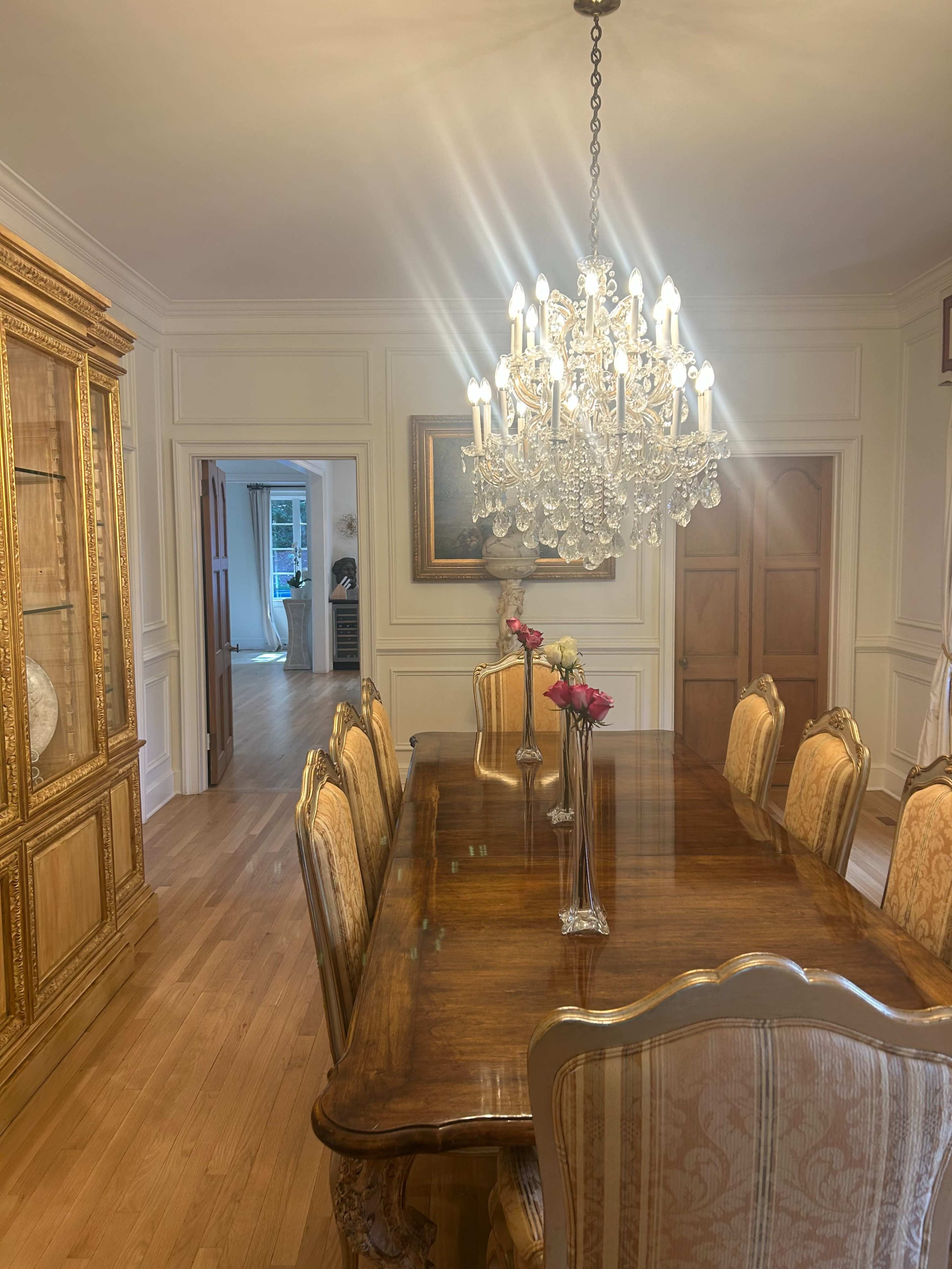 A long wooden dining table is surrounded by upholstered chairs, with a crystal chandelier hanging above and a cabinet displaying decorative items in the background.
