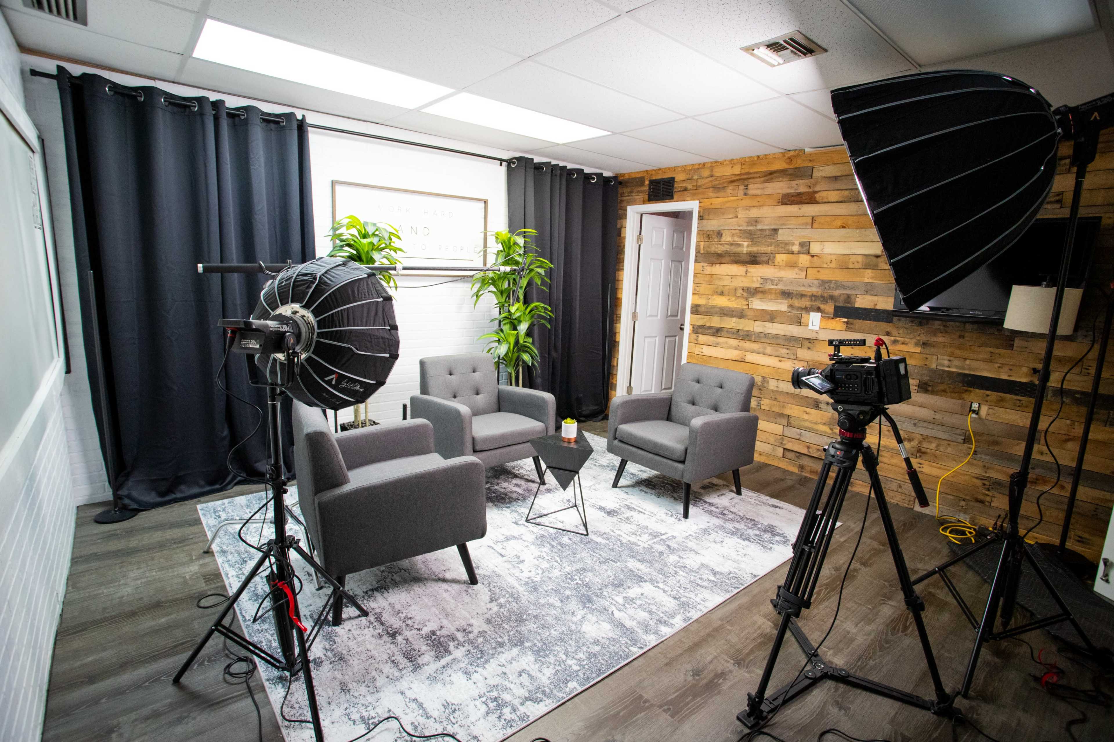 The image shows a well-lit studio setup featuring two gray chairs, a small black table, and equipment including a camera and lights, with a wooden accent wall in the background.