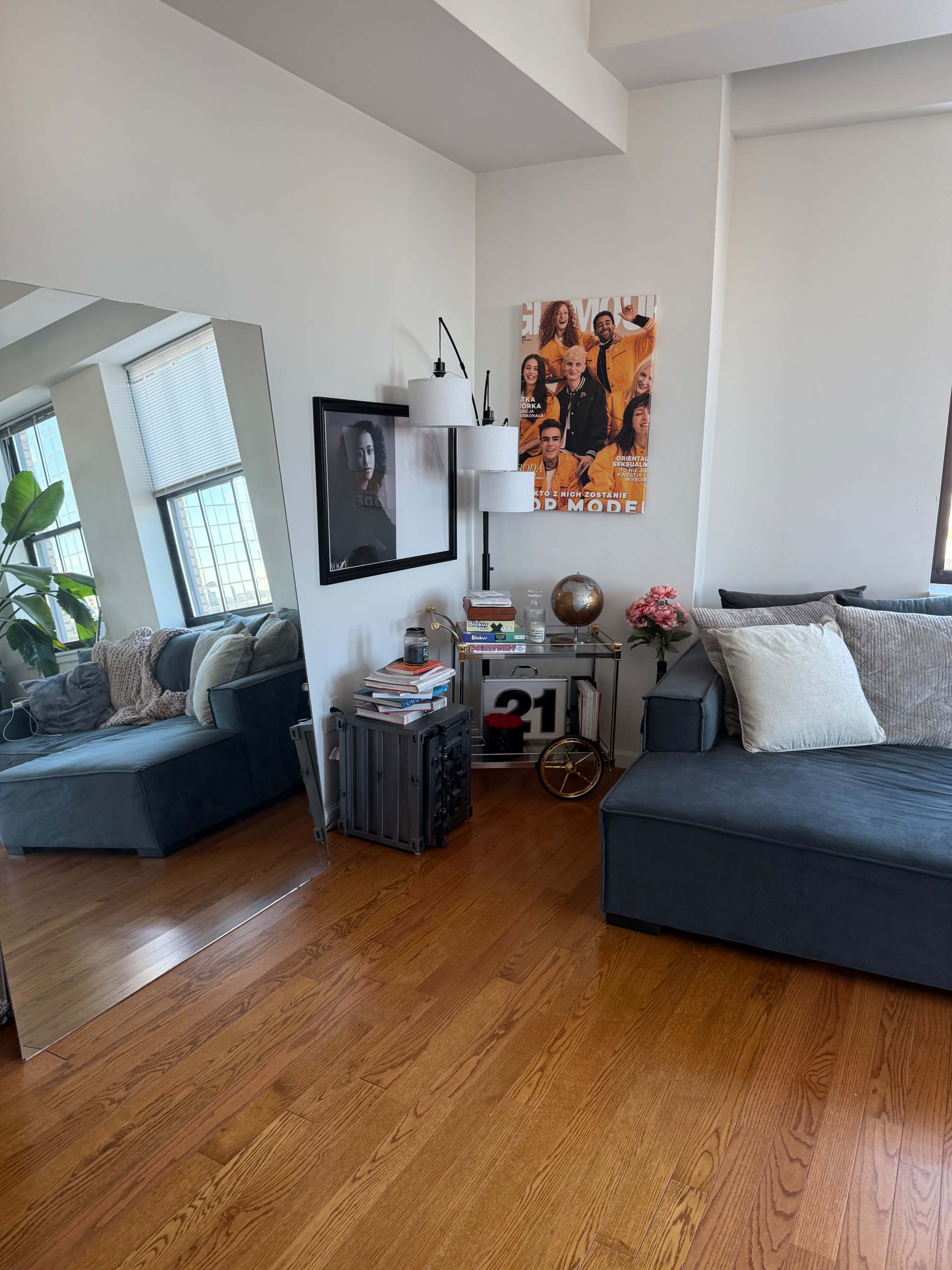 The image shows a cozy living room corner with a blue sofa, a mirrored wall, a small cart with books and a globe, and a large poster on the wall.