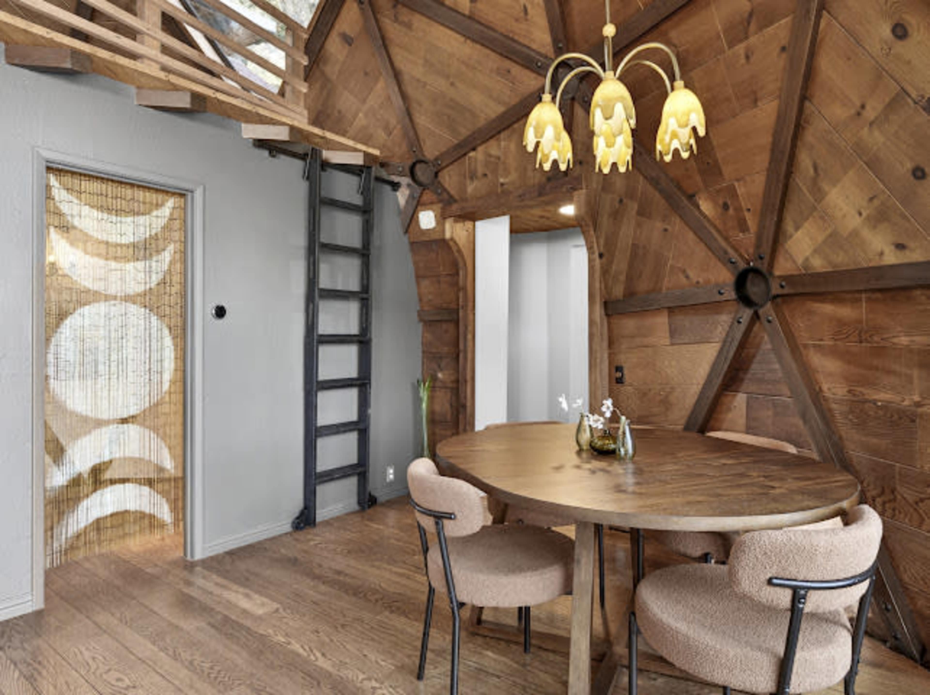 A circular wooden dining table is surrounded by four upholstered chairs in a room with wooden walls and a distinctive ceiling design, featuring a ladder leading to an upper level.