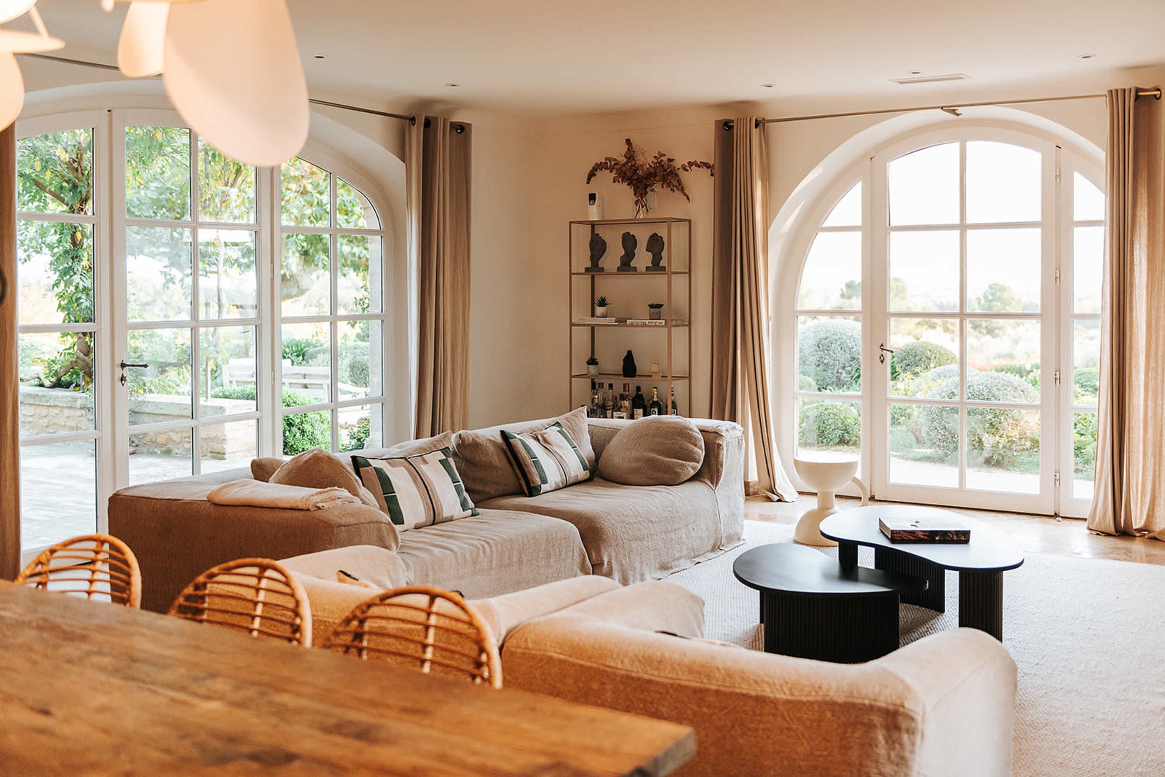 The image shows a spacious living room with large arched windows, a light-colored sectional sofa, a round coffee table, and a wooden dining table in the foreground.
