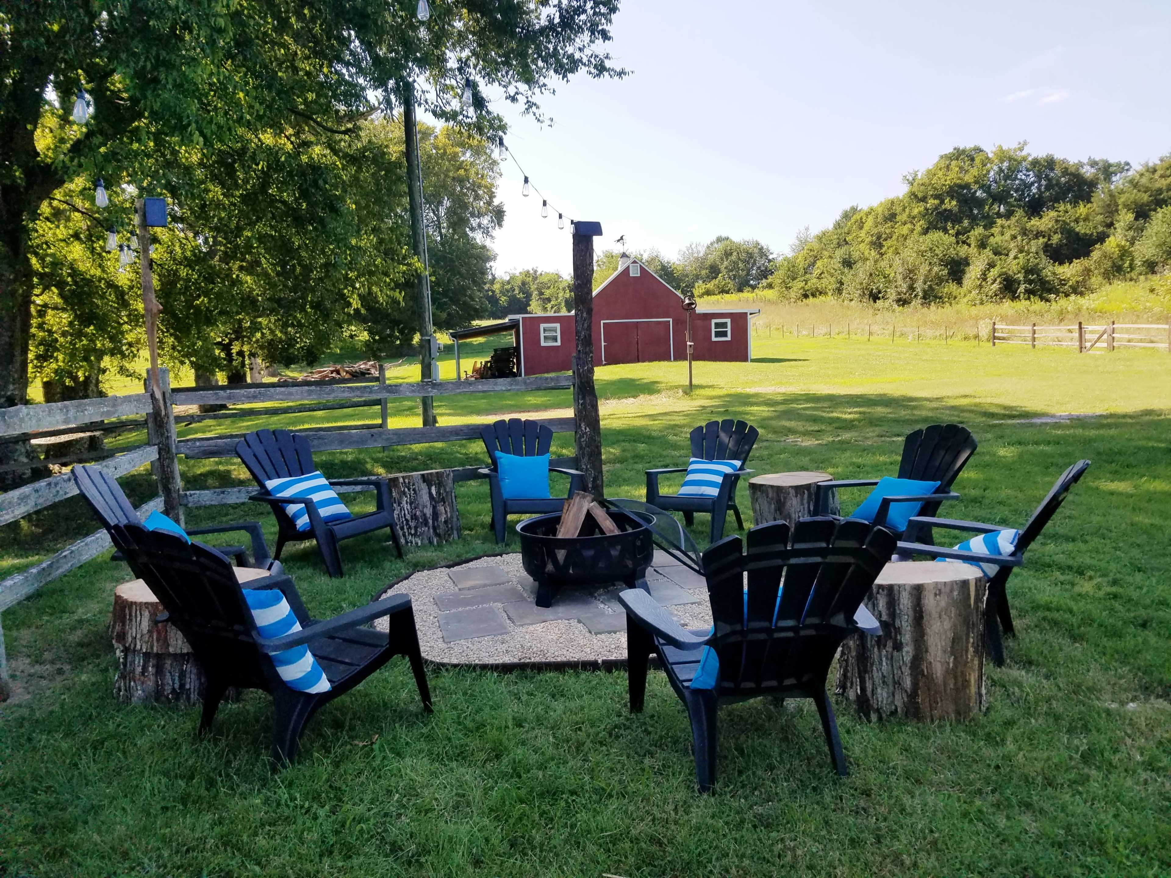 A circular arrangement of black Adirondack chairs around a stone fire pit in a grassy area, with a red barn and trees in the background.