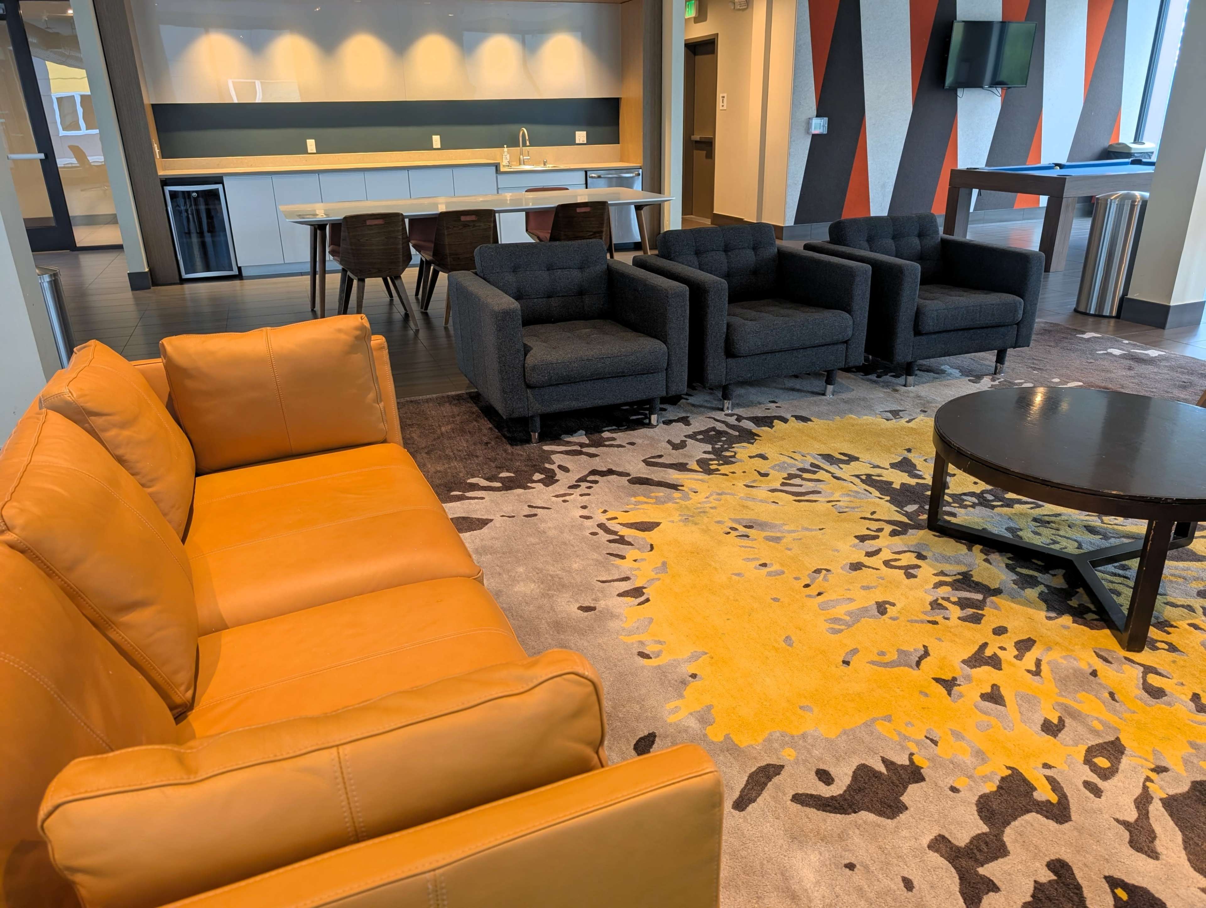 The image shows a modern lounge area with a tan leather sofa, black armchairs, a round coffee table, and a patterned rug on the floor.
