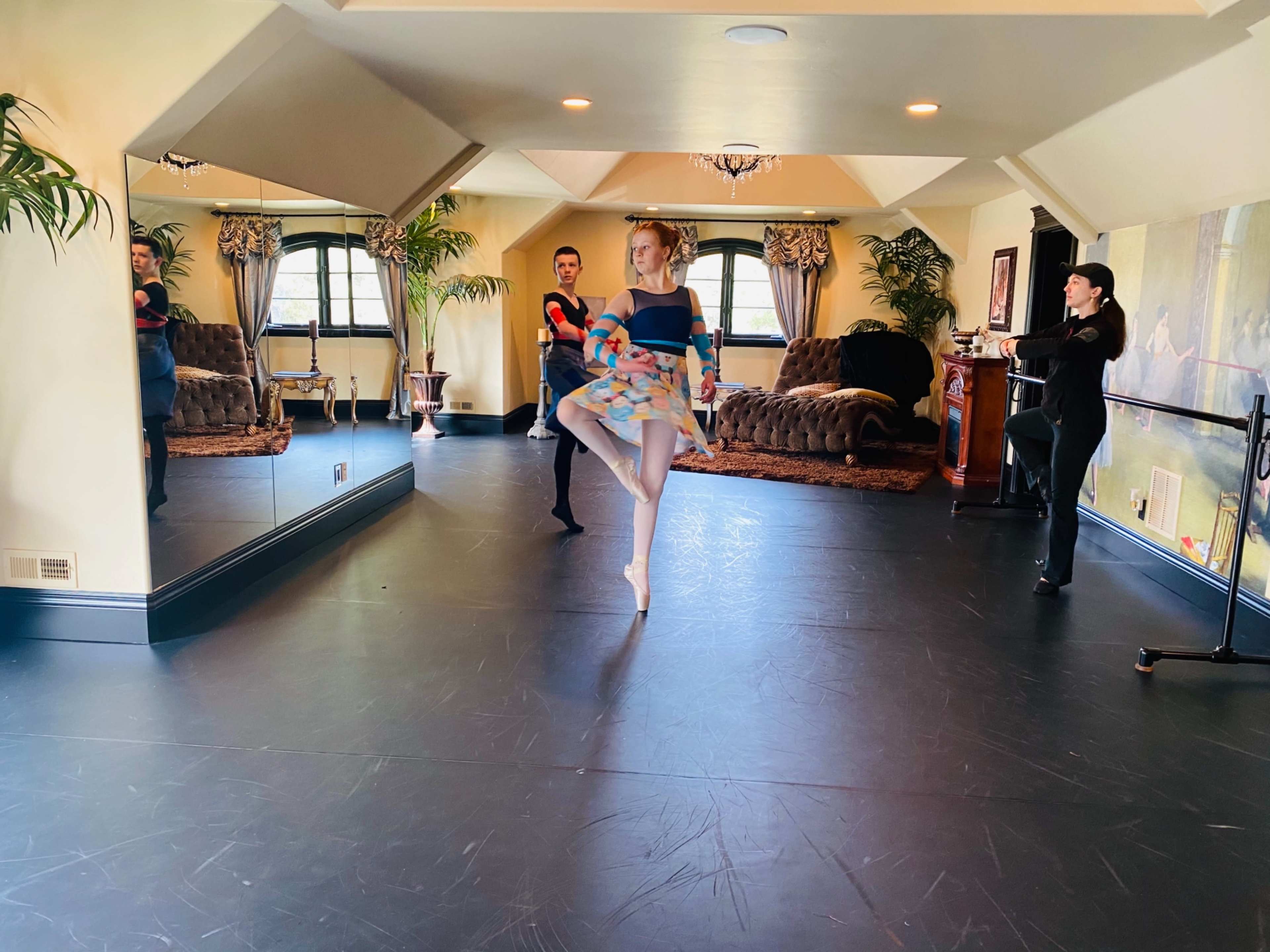 A ballet dancer performs a pose in a studio while two instructors observe in the background.