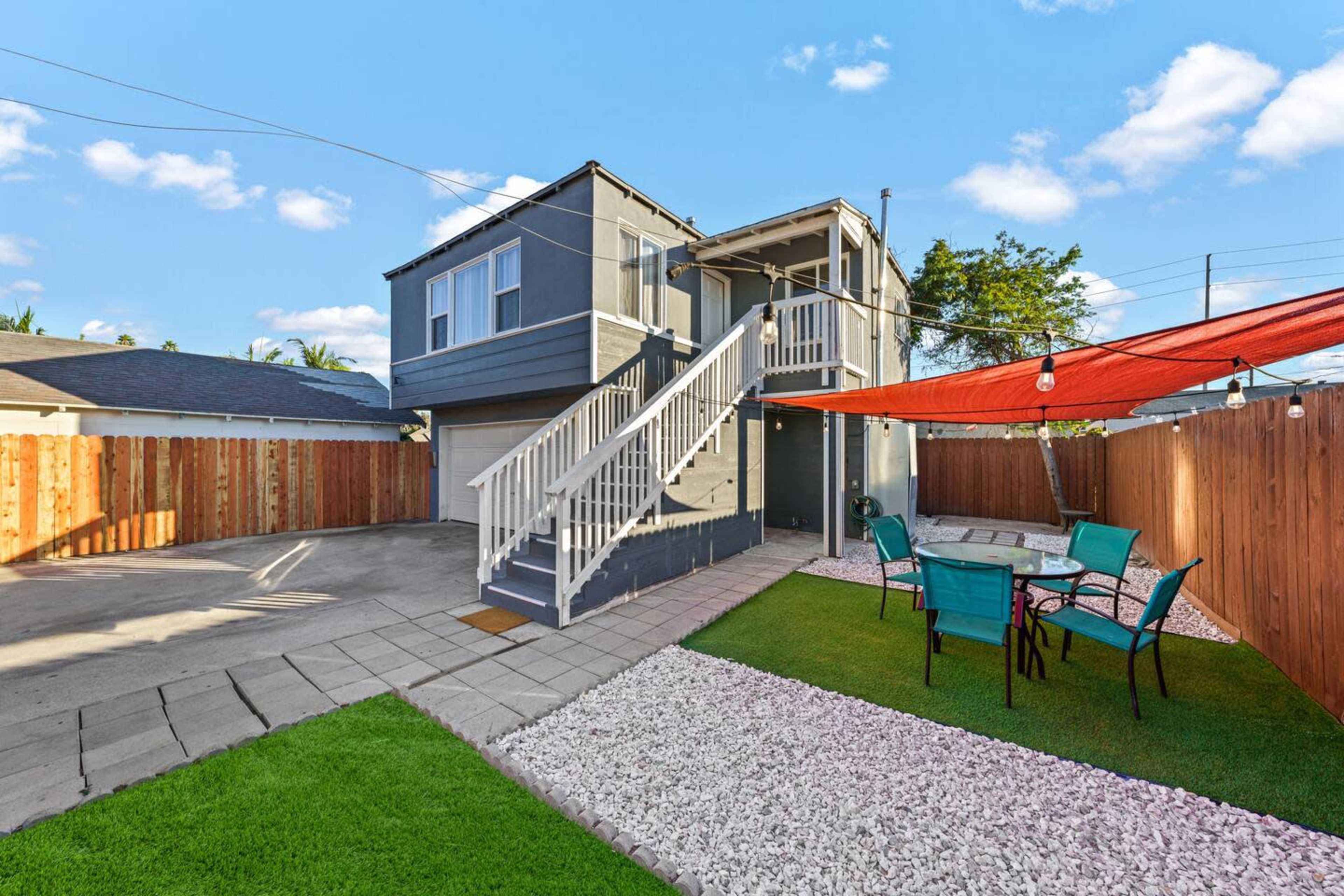 The image shows a two-story gray house with a staircase leading to the upper level, set in a yard featuring a patio area with outdoor chairs and a gravel pathway, surrounded by wooden fencing.