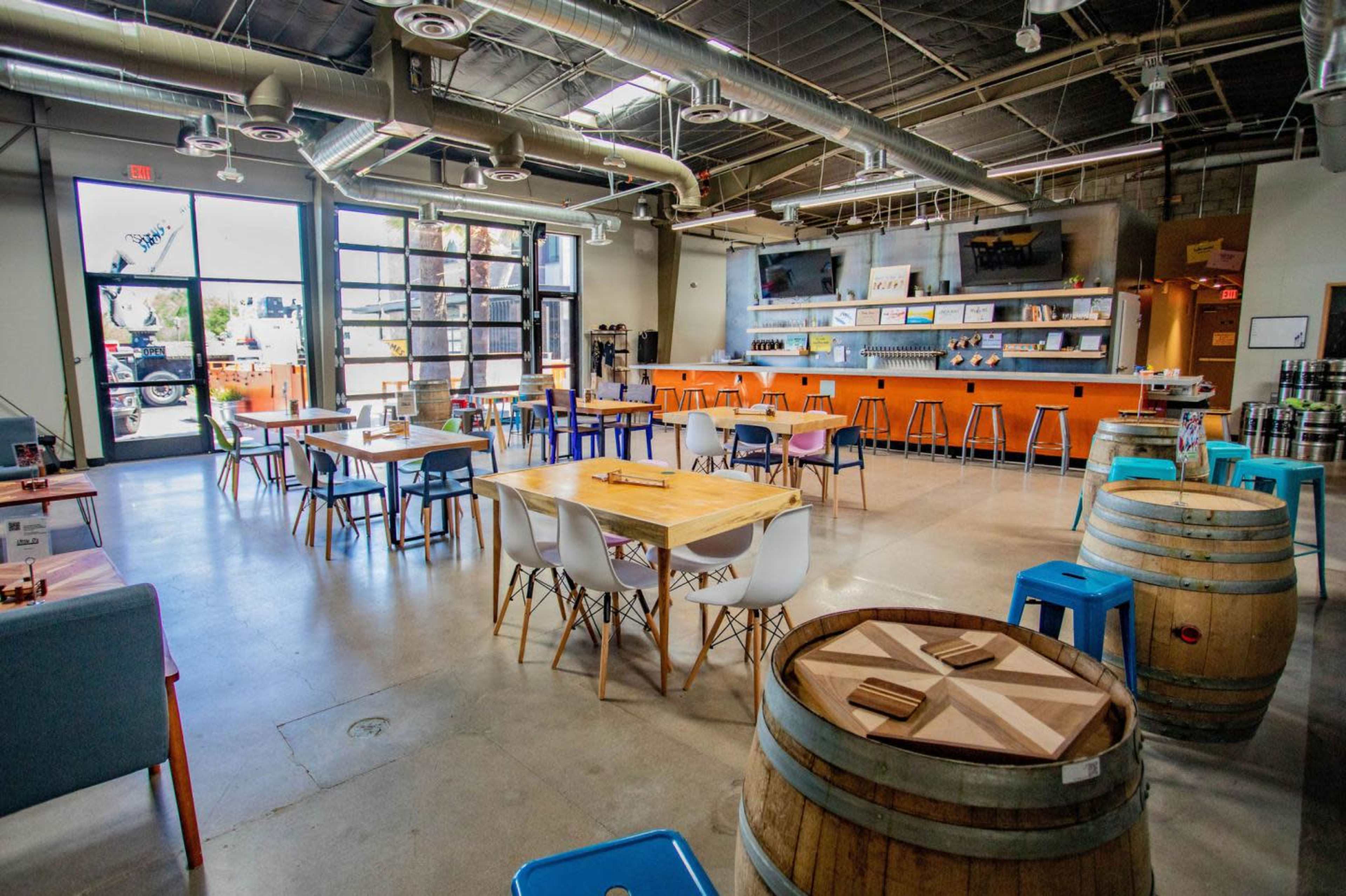 The image shows an industrial-style interior of a spacious bar or restaurant with various seating options, including wooden tables, colorful stools, and barrels, along with a counter against a wall featuring screens.