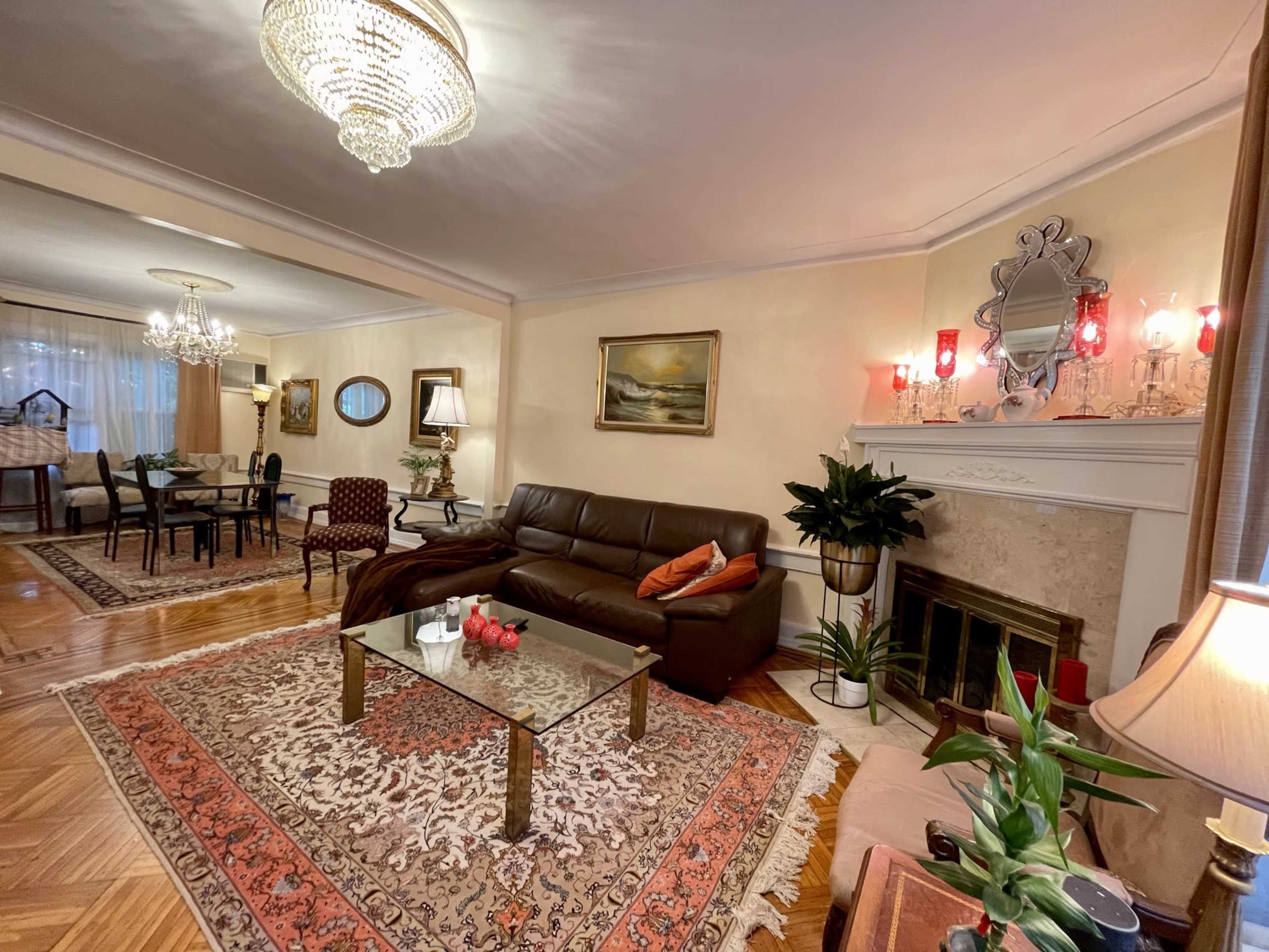 A cozy living room featuring a brown sofa, a glass coffee table, a fireplace, and decorative plants, with a dining area visible in the background.