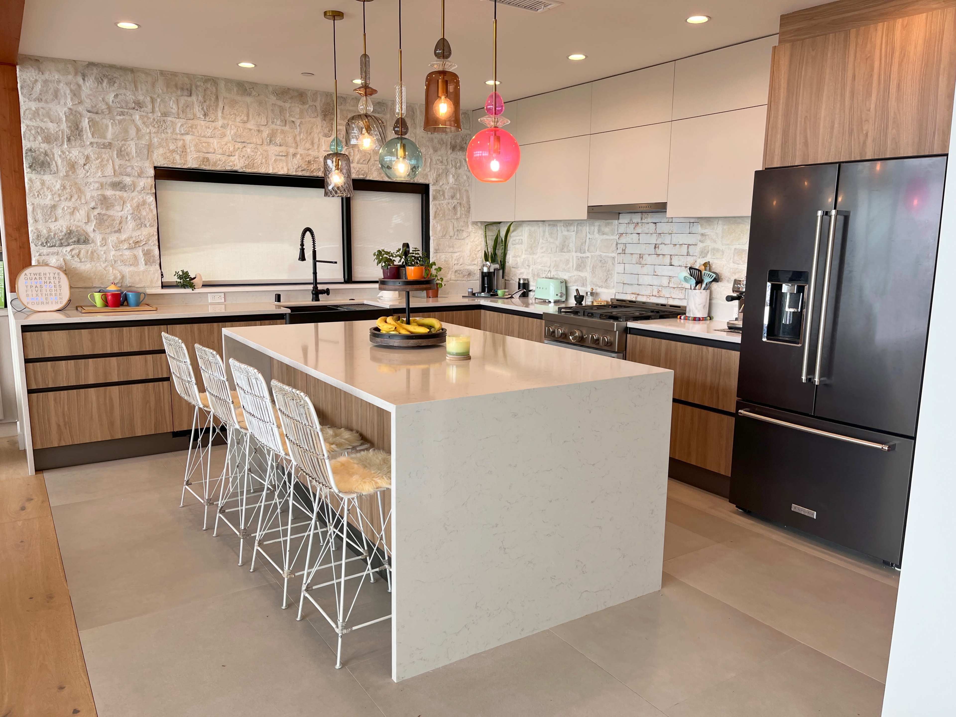 A modern kitchen features a large island with bar seating, colorful pendant lights, and a stone wall backdrop.