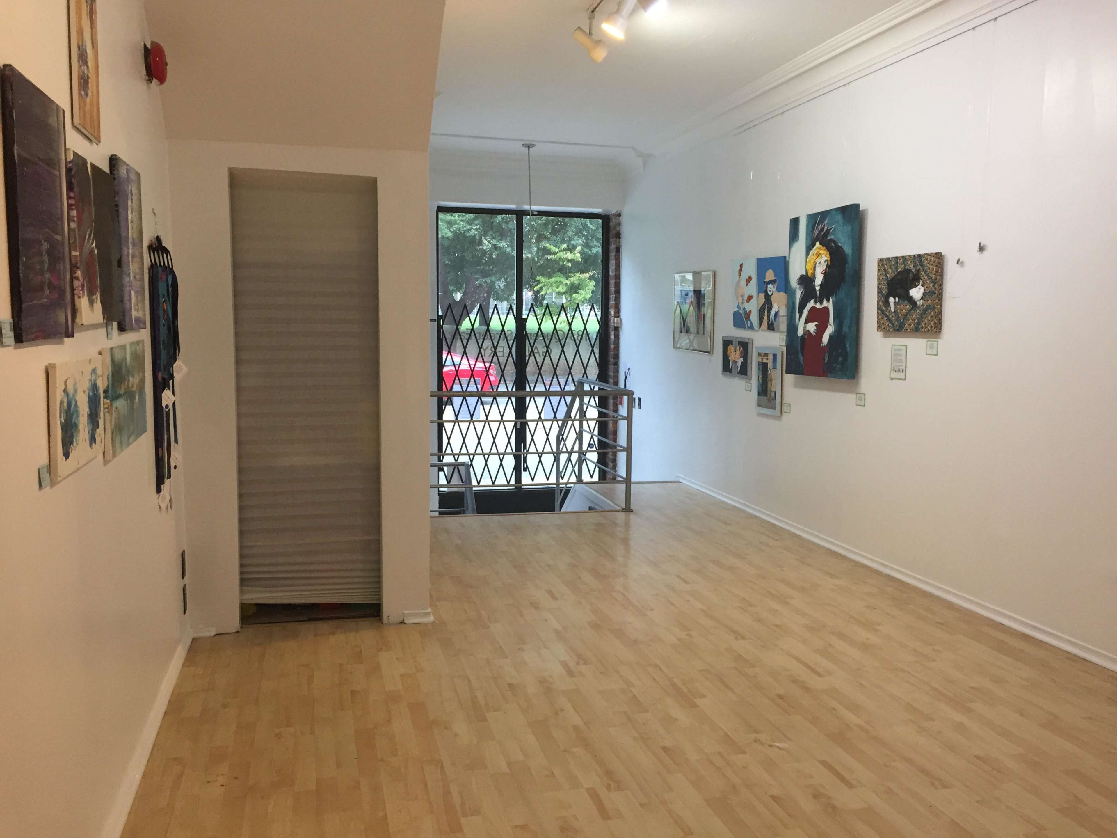 A gallery space with wood flooring, featuring various artworks on the walls and a locked gate leading to the outside.