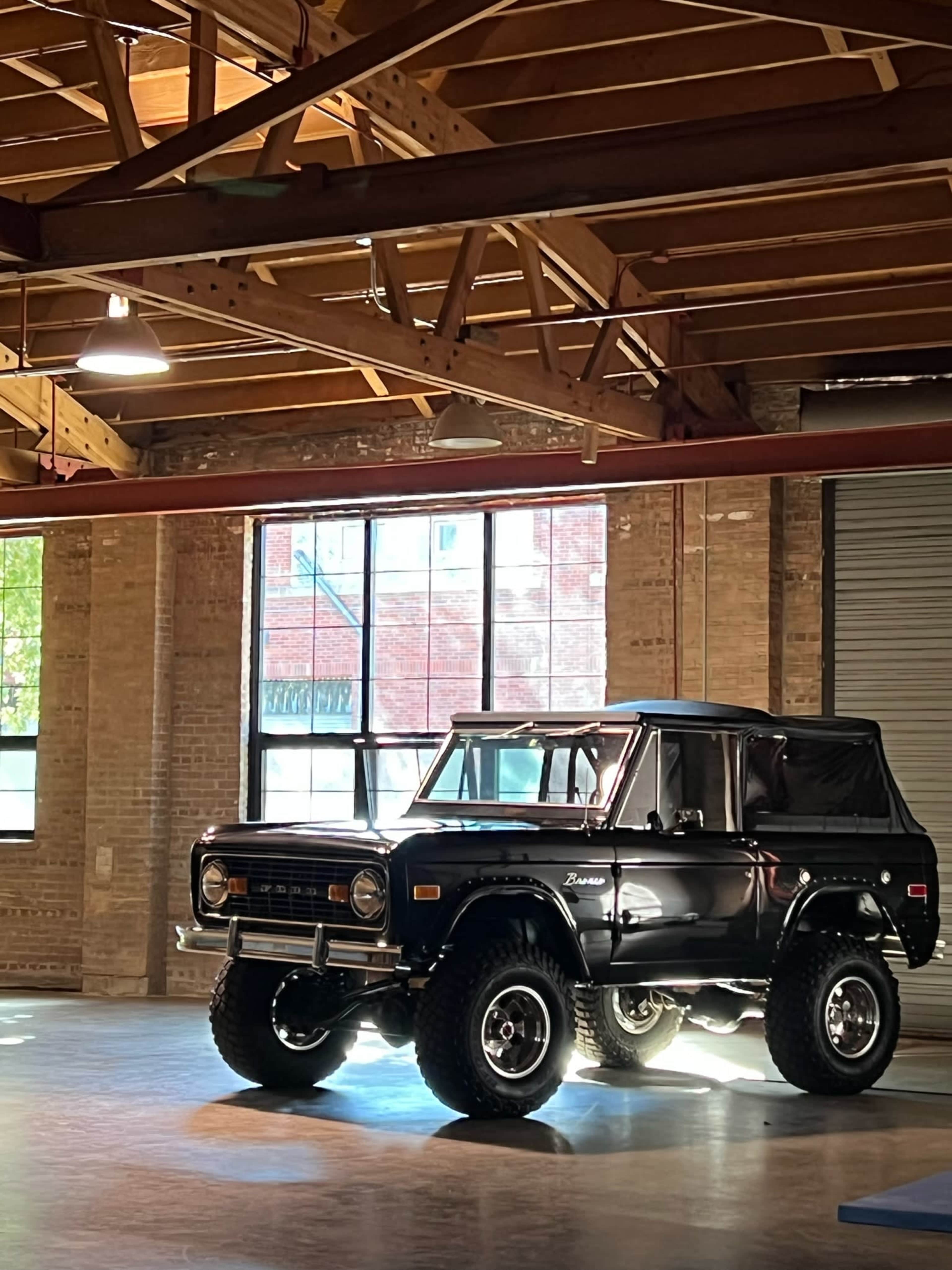 A vintage black Bronco is parked on a polished floor in a spacious warehouse with large windows.