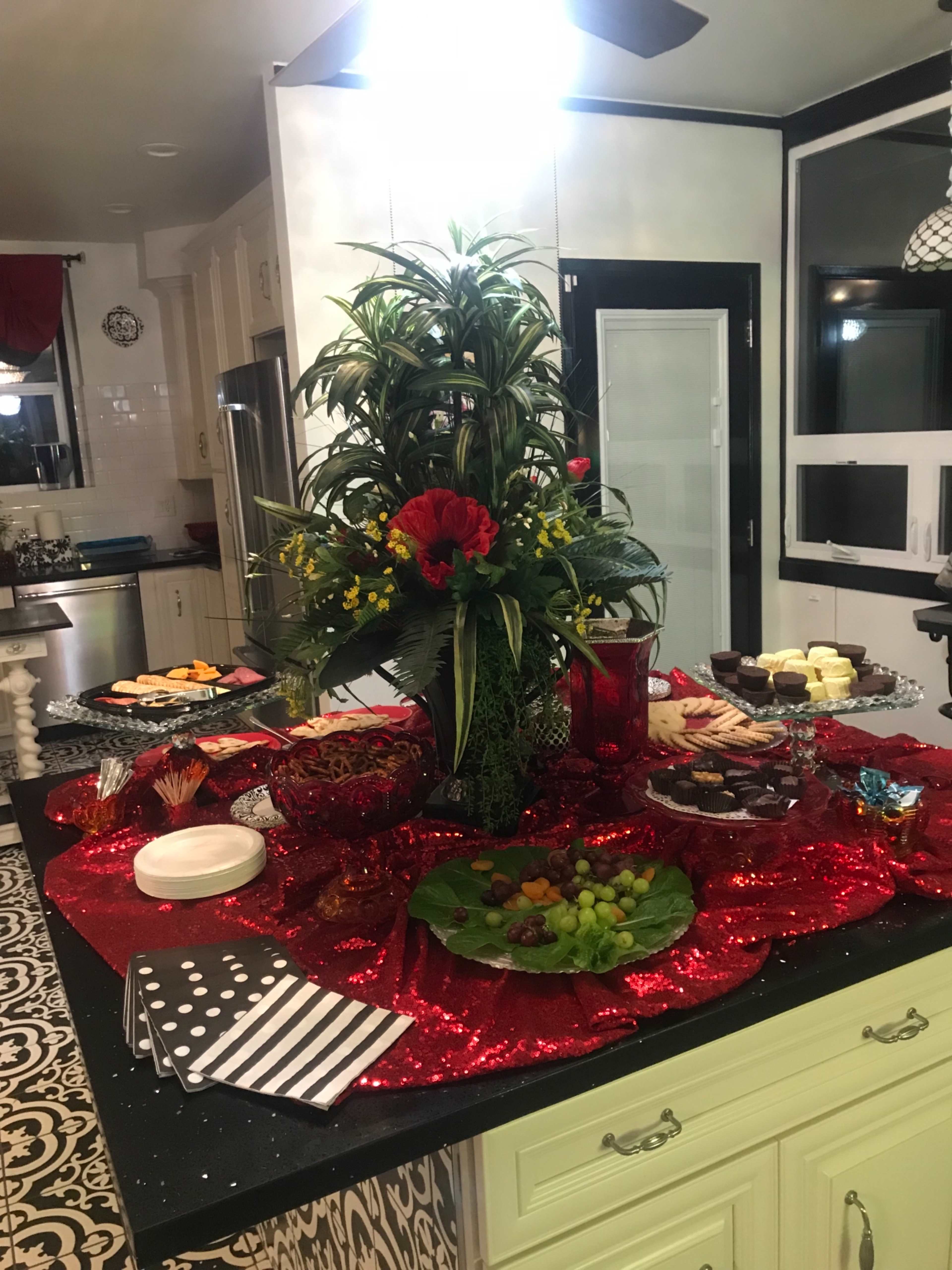 The image features a decorative kitchen island adorned with a large floral centerpiece and various dishes of appetizers and desserts arranged on a red sparkly tablecloth.