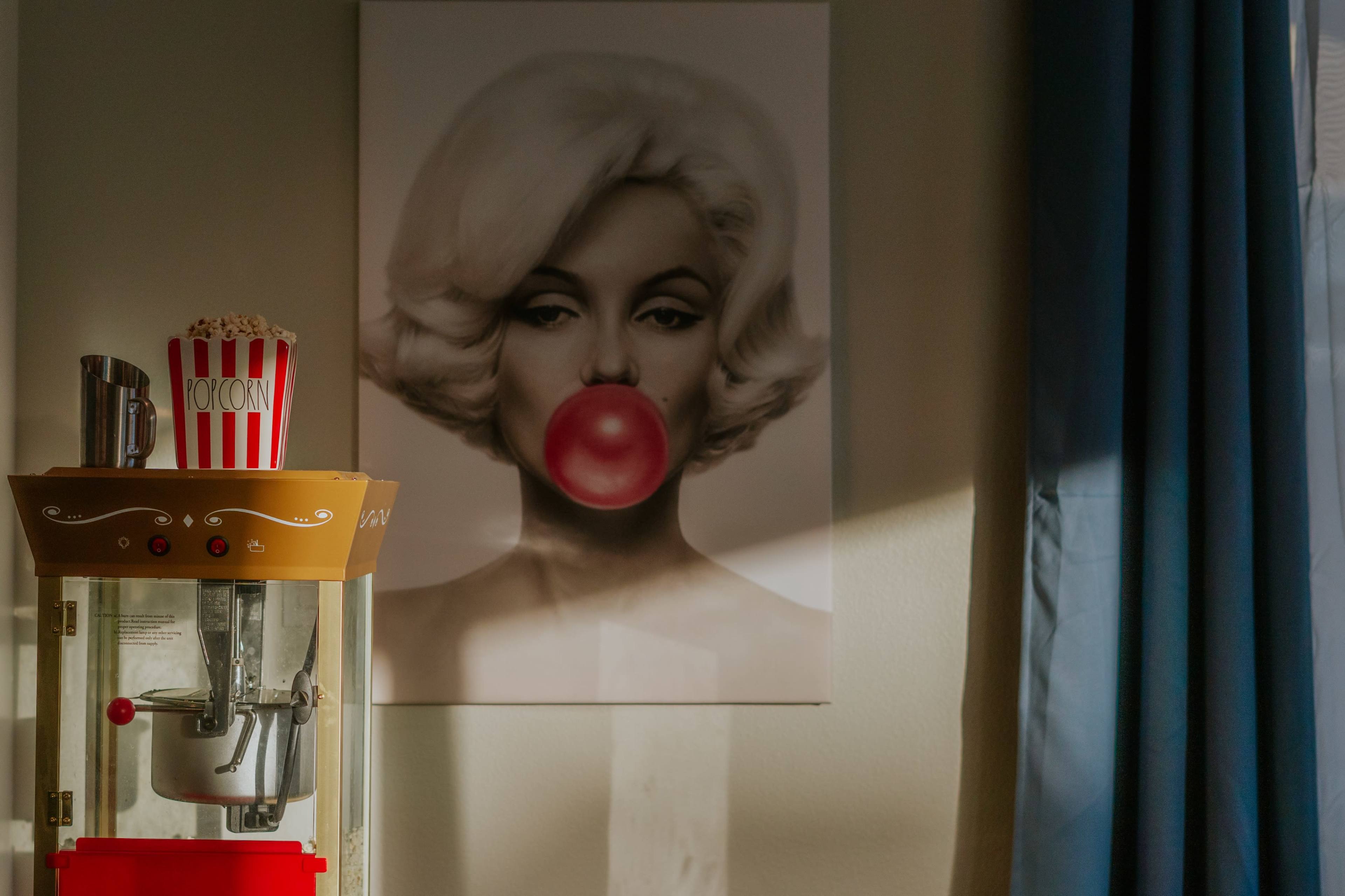 A vintage popcorn machine sits next to a poster of a woman blowing a bubblegum bubble, with blue curtains in the background.