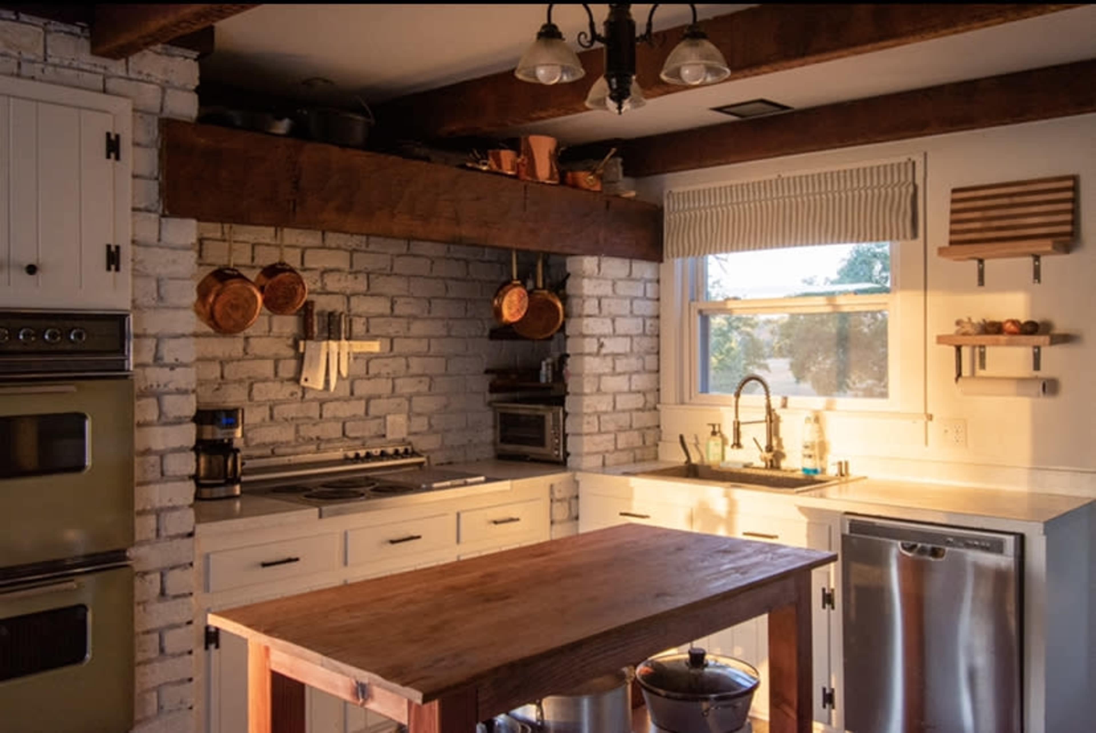 A kitchen with white brick walls, wooden beams, and a central wooden island with various kitchen appliances and utensils displayed.