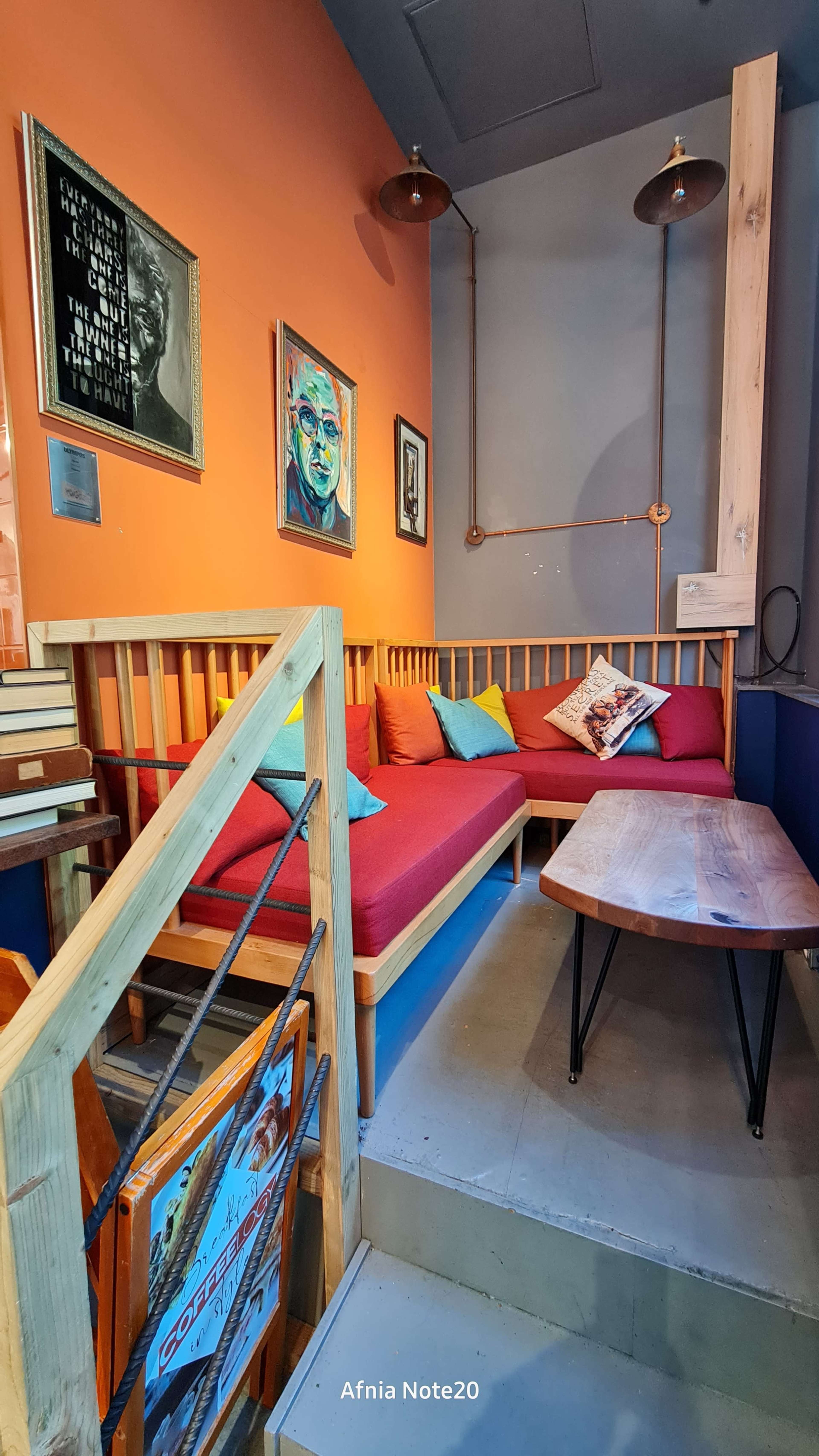 The image shows a small seating area in a café featuring a wooden bench with colorful cushions, a wooden table, and framed artwork on the walls.