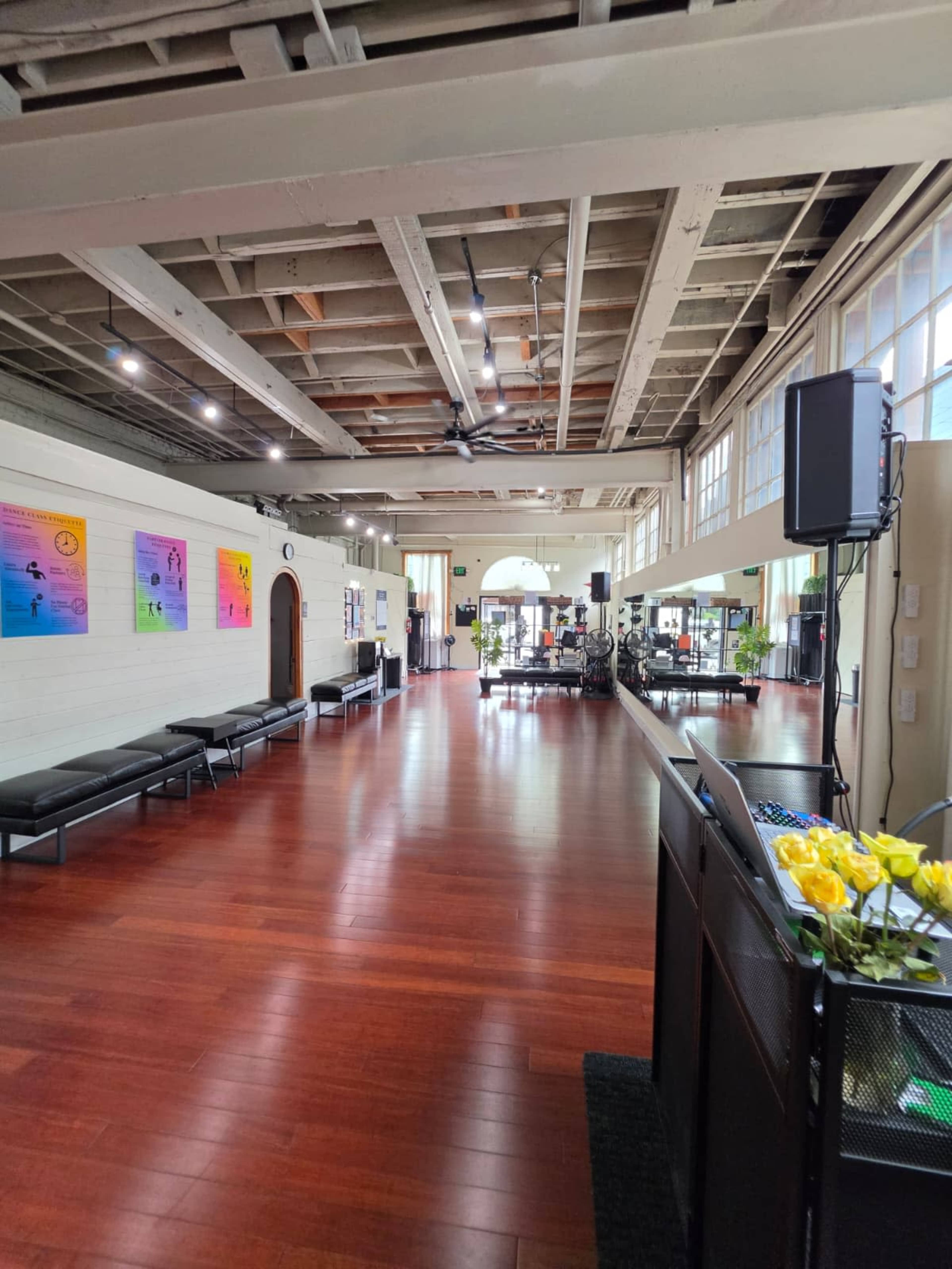 The image shows the interior of a spacious fitness studio with wooden flooring, bright windows, and various exercise equipment along the walls.