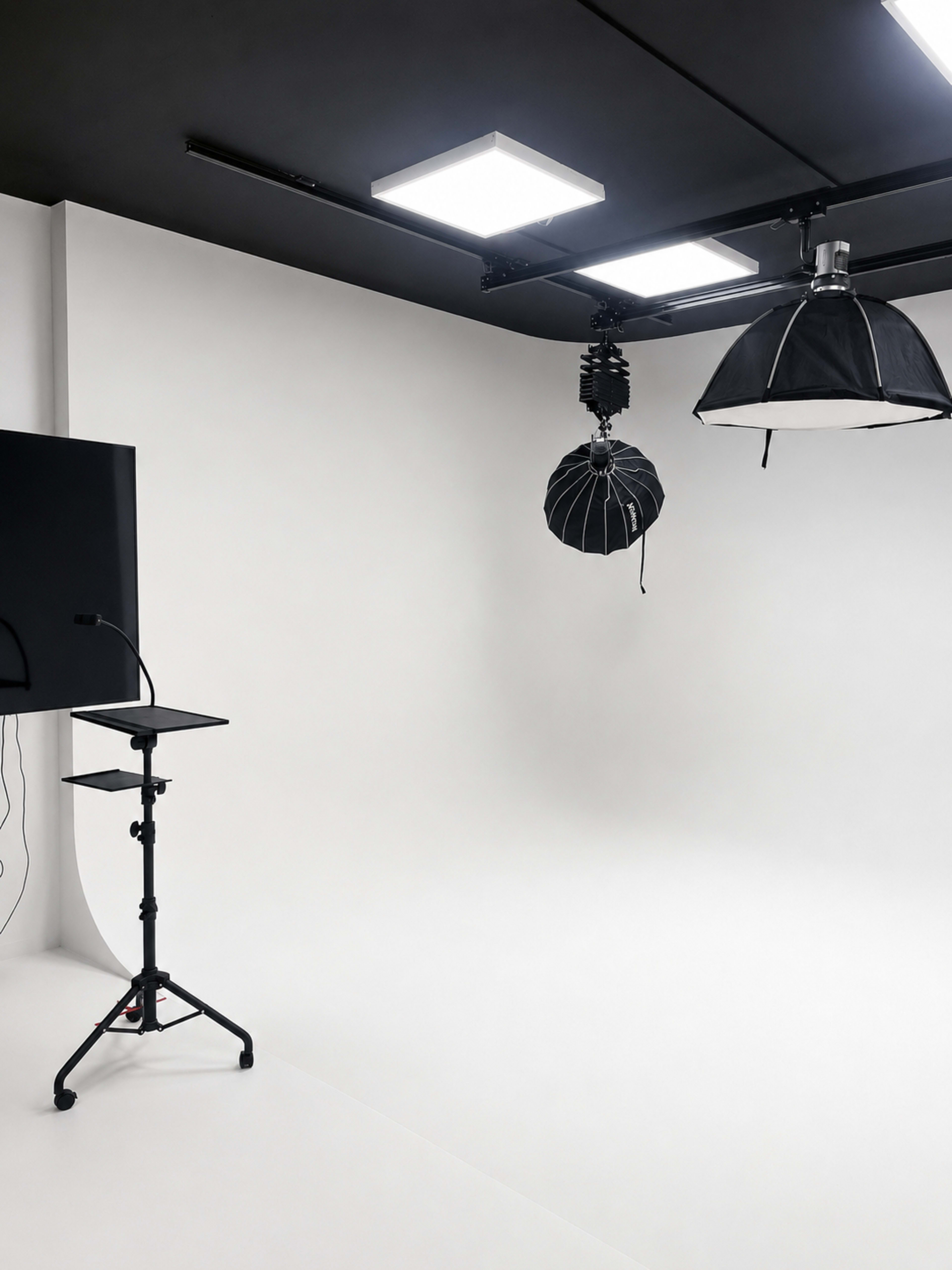 The image shows a photo studio with a stark white backdrop, lighting setups, and a mounted monitor.