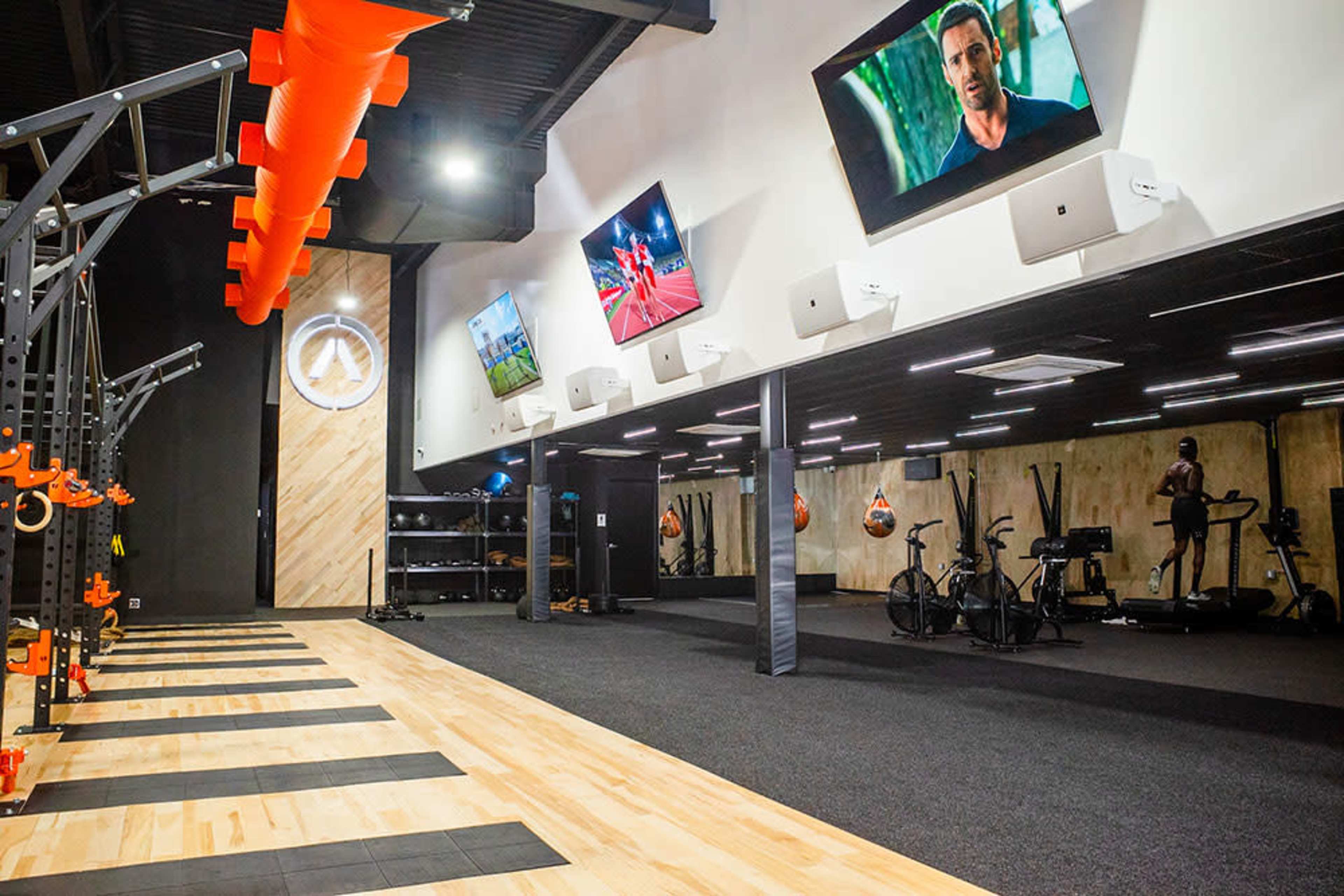 A modern fitness gym with various workout stations, including exercise bikes and a section for strength training, illuminated by overhead lights and featuring multiple screens on the walls.