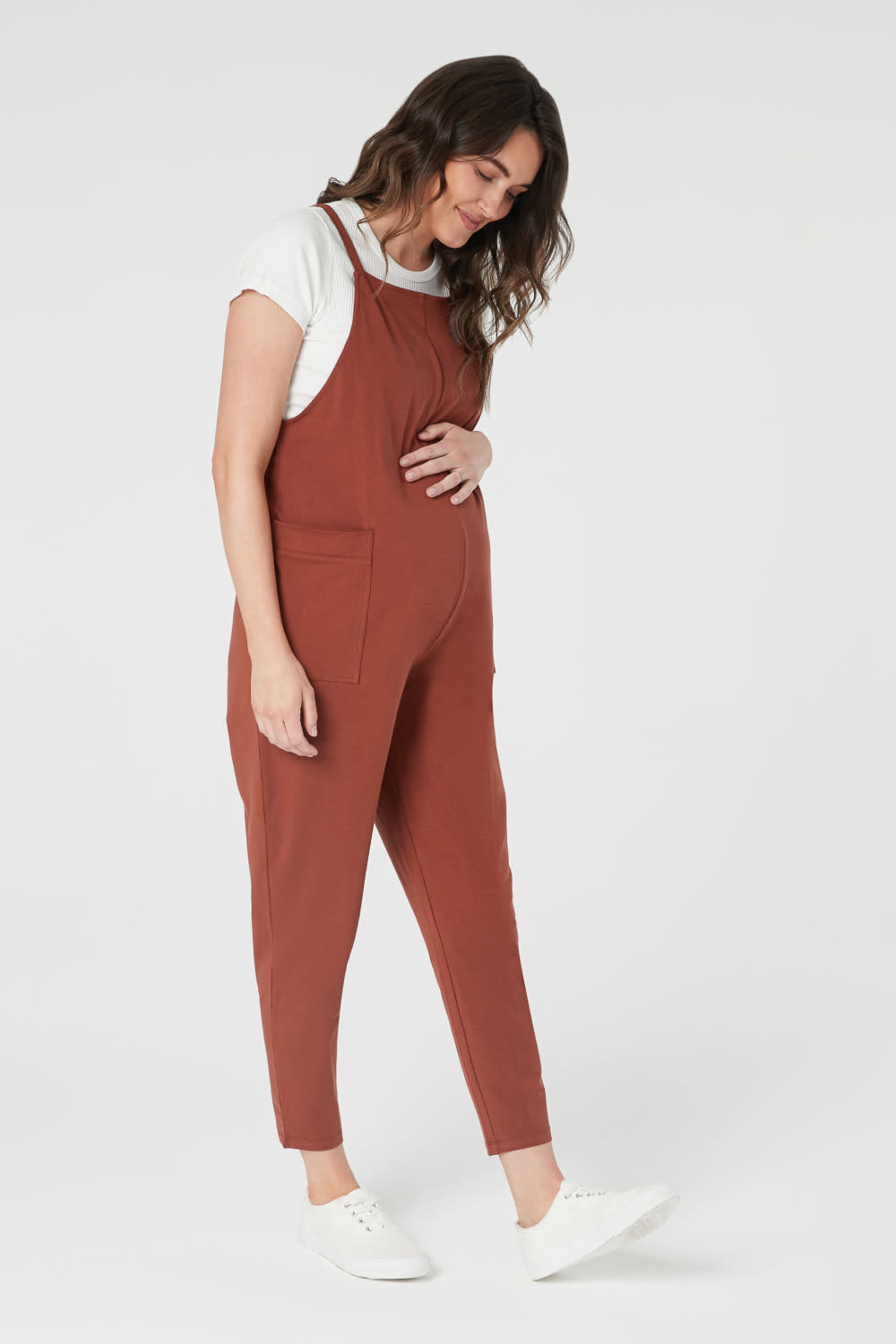 A woman wearing a rust-colored jumpsuit and a white t-shirt stands with her hand on her stomach.