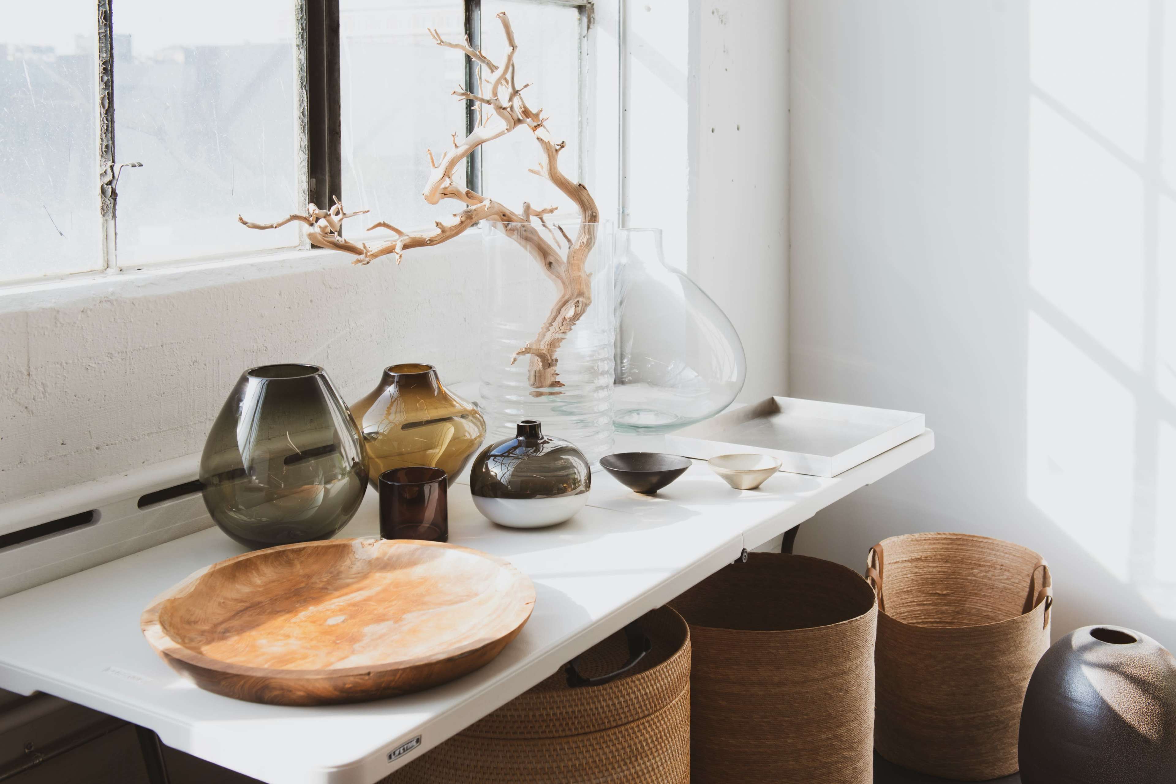 A collection of glass vases, bowls, and woven baskets is arranged on a table near a window.