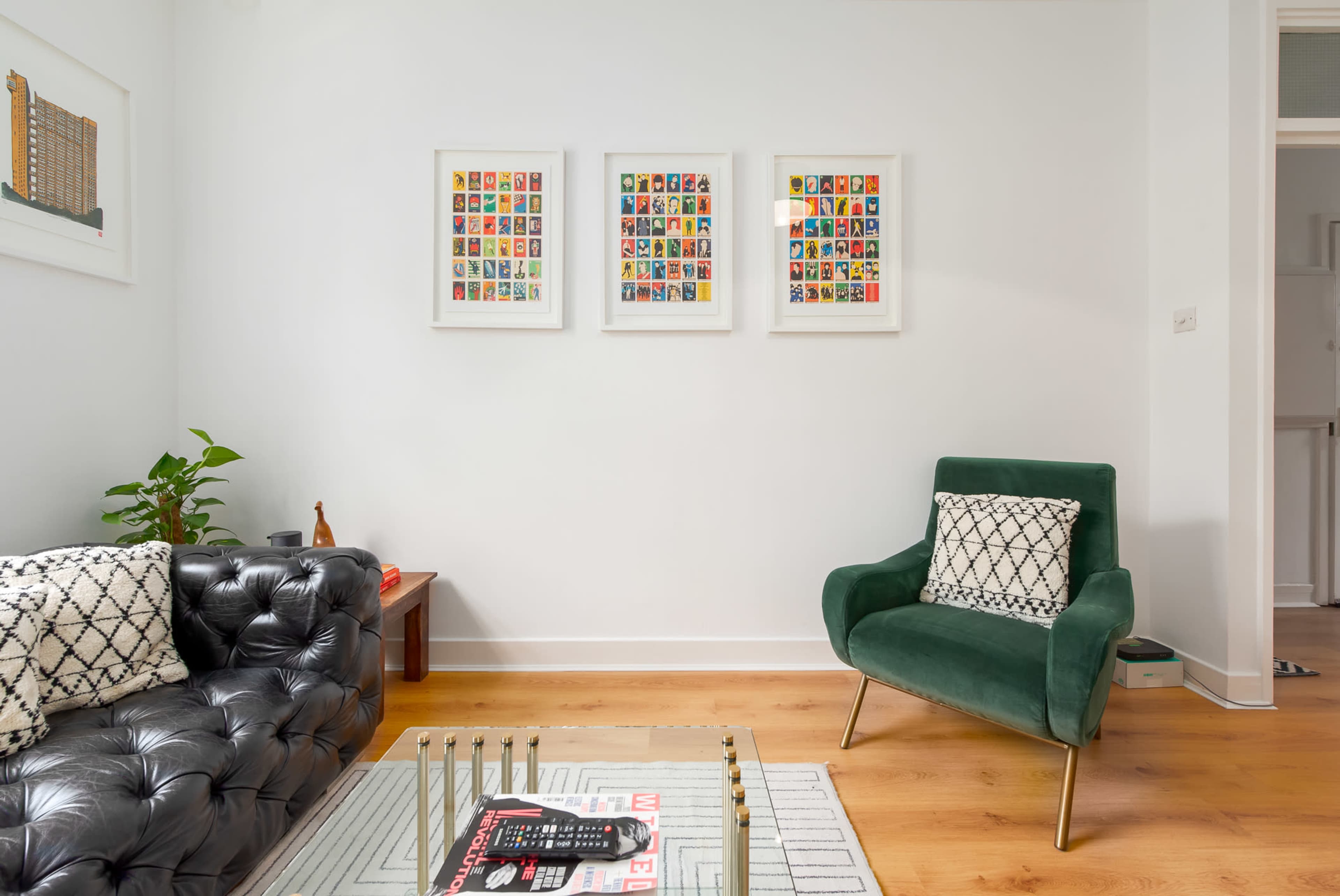 A modern living room featuring a black leather sofa, a green armchair, and three framed pieces of colorful artwork on the white wall.
