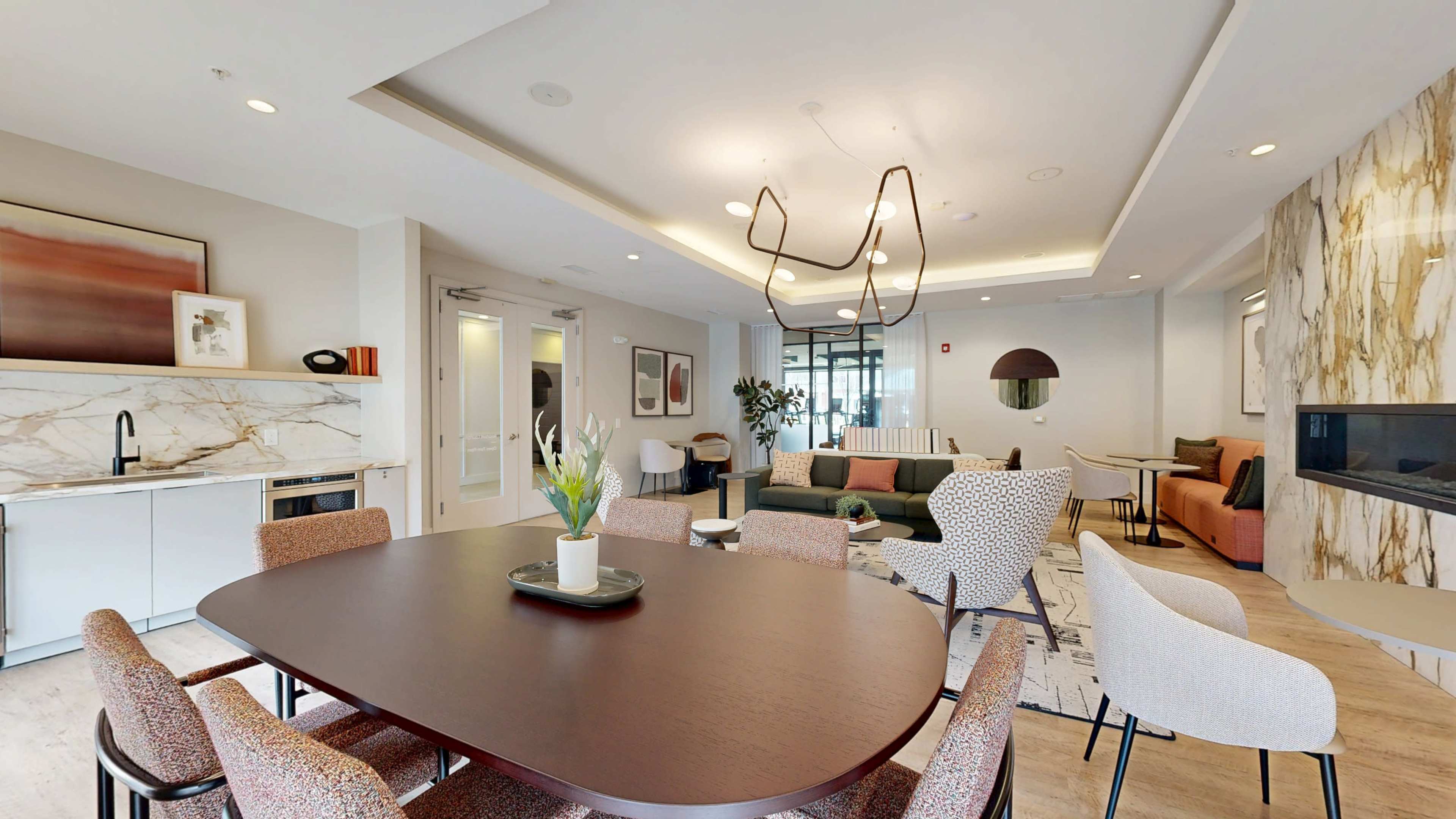 A modern communal lounge area features a large oval dining table surrounded by upholstered chairs, with a cozy seating arrangement in the background and decorative wall art.