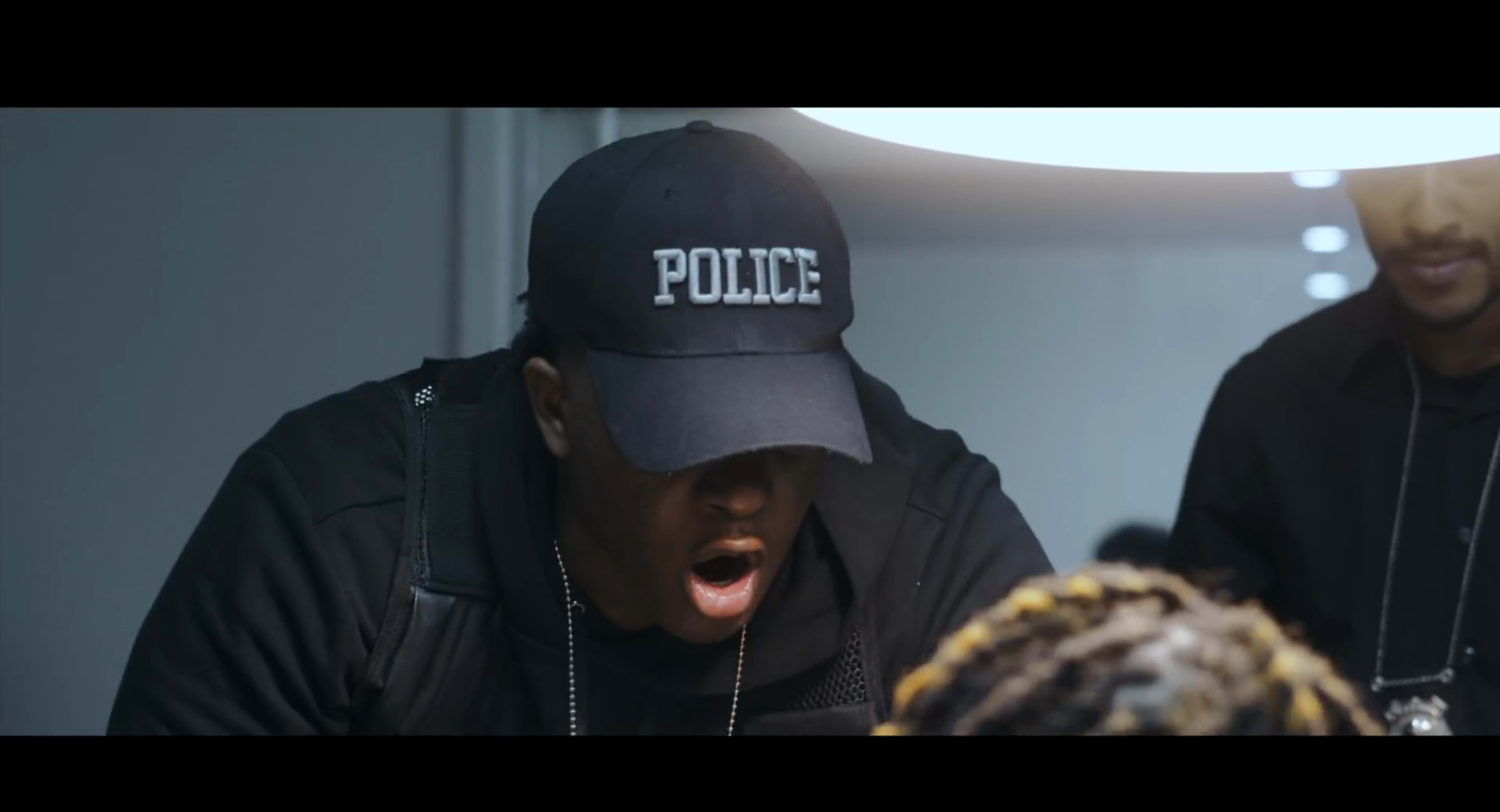 A police officer wearing a cap with "POLICE" written on it engages in a serious conversation with another person in an interrogation room.