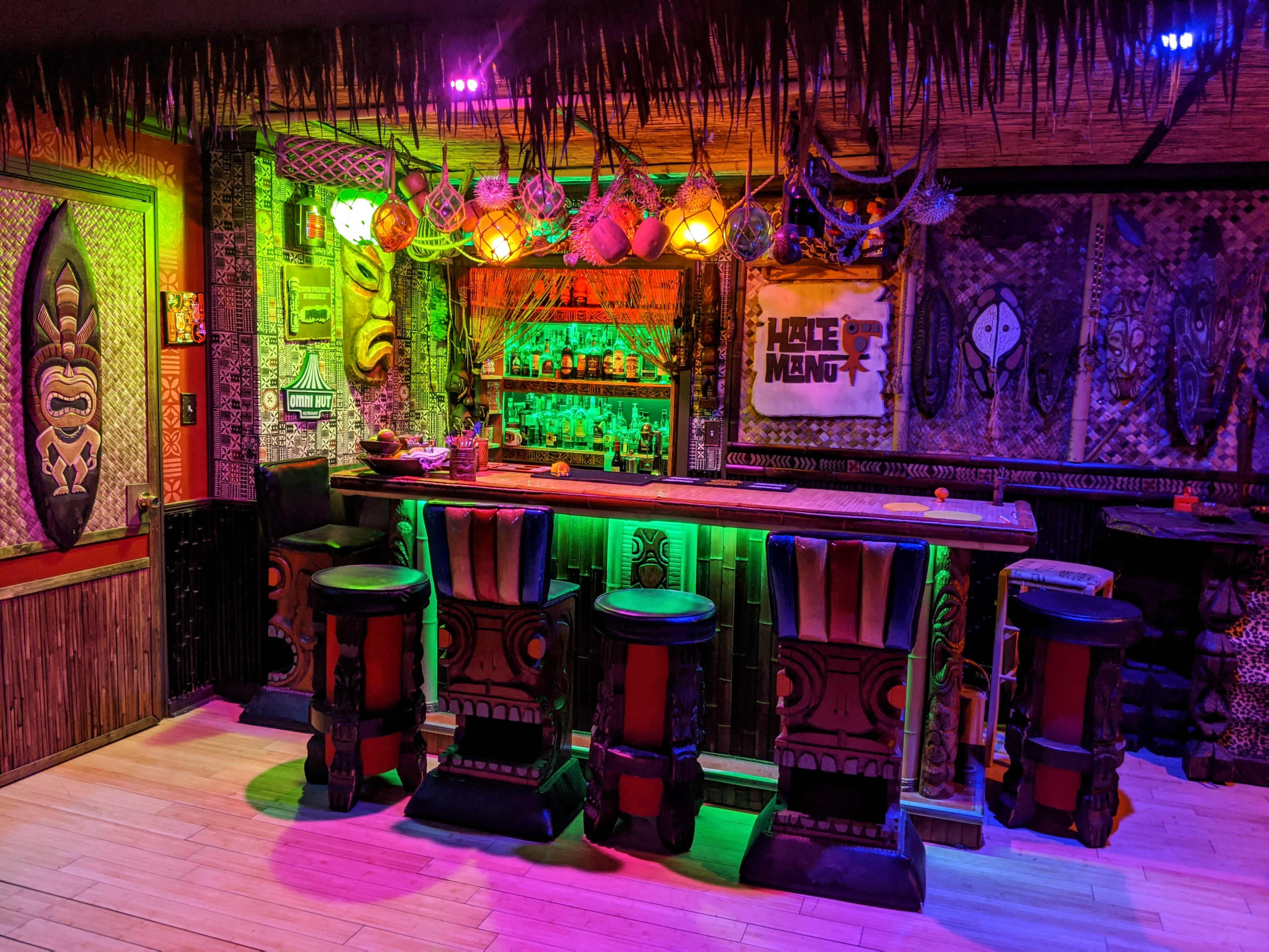 The image shows a vibrant tiki bar featuring colorful lighting, a wooden bar with decorative stools, and tropical-themed decor on the walls.