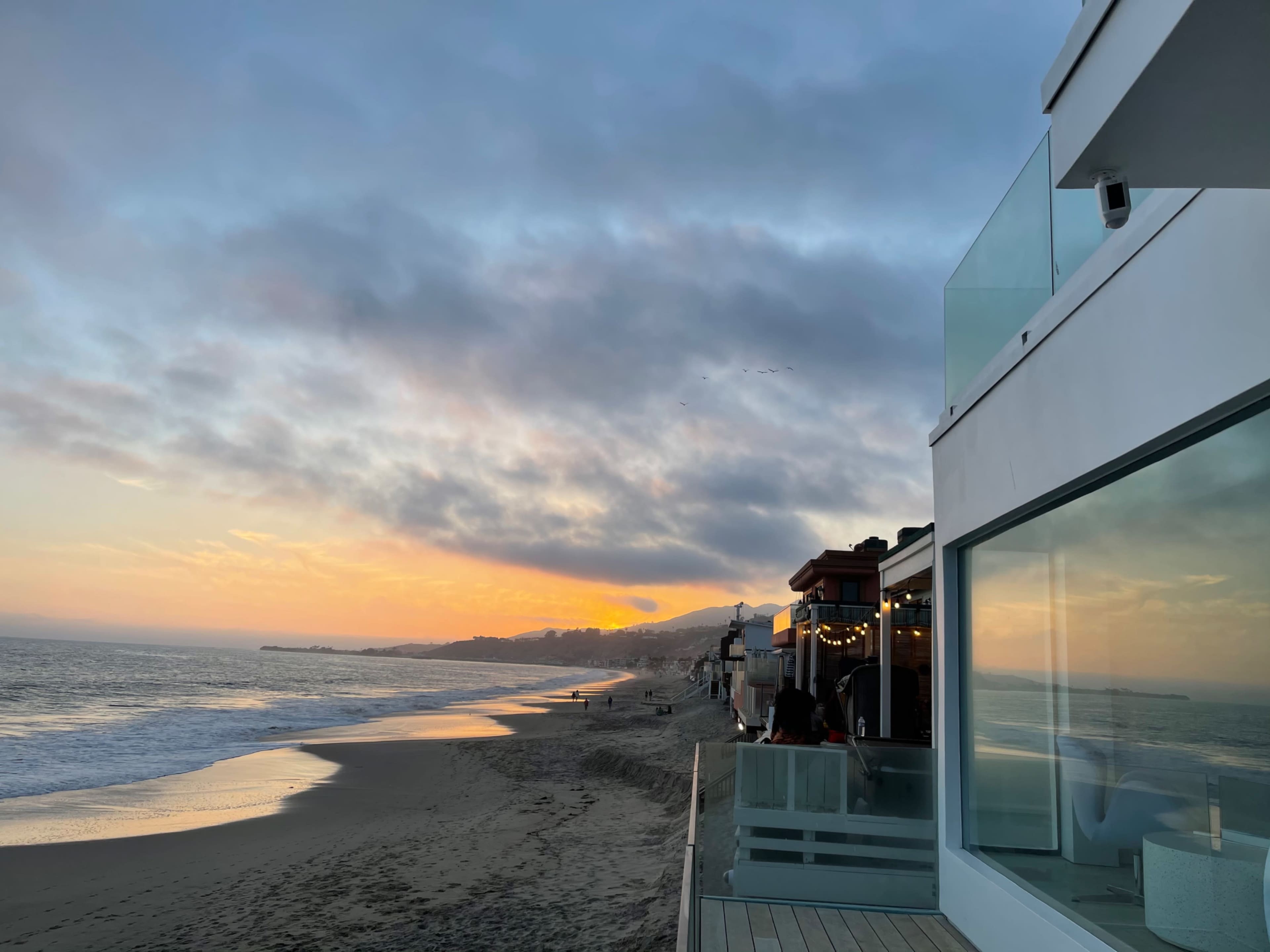 The image shows a beach at sunset with waves lapping at the shore and a modern building overlooking the scene.
