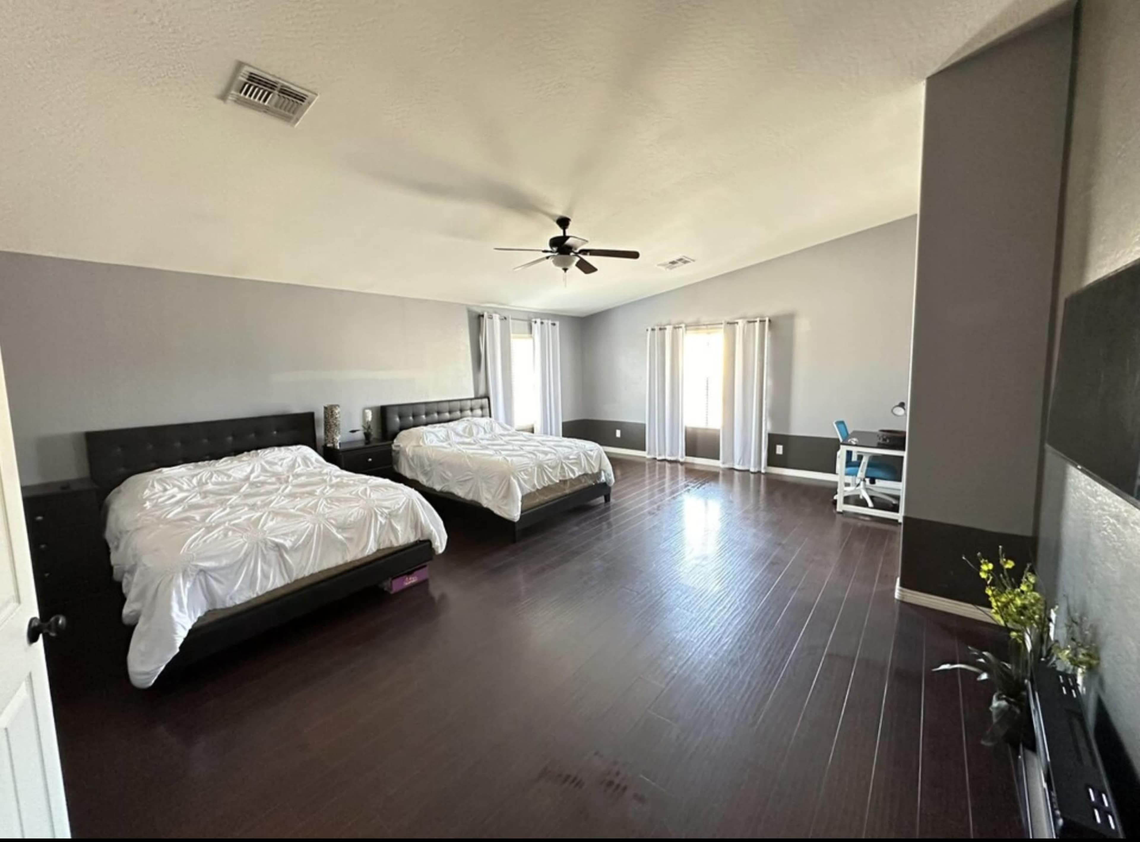 The image shows a spacious bedroom with two large beds, wooden flooring, and a desk against a wall with windows covered by sheer curtains.