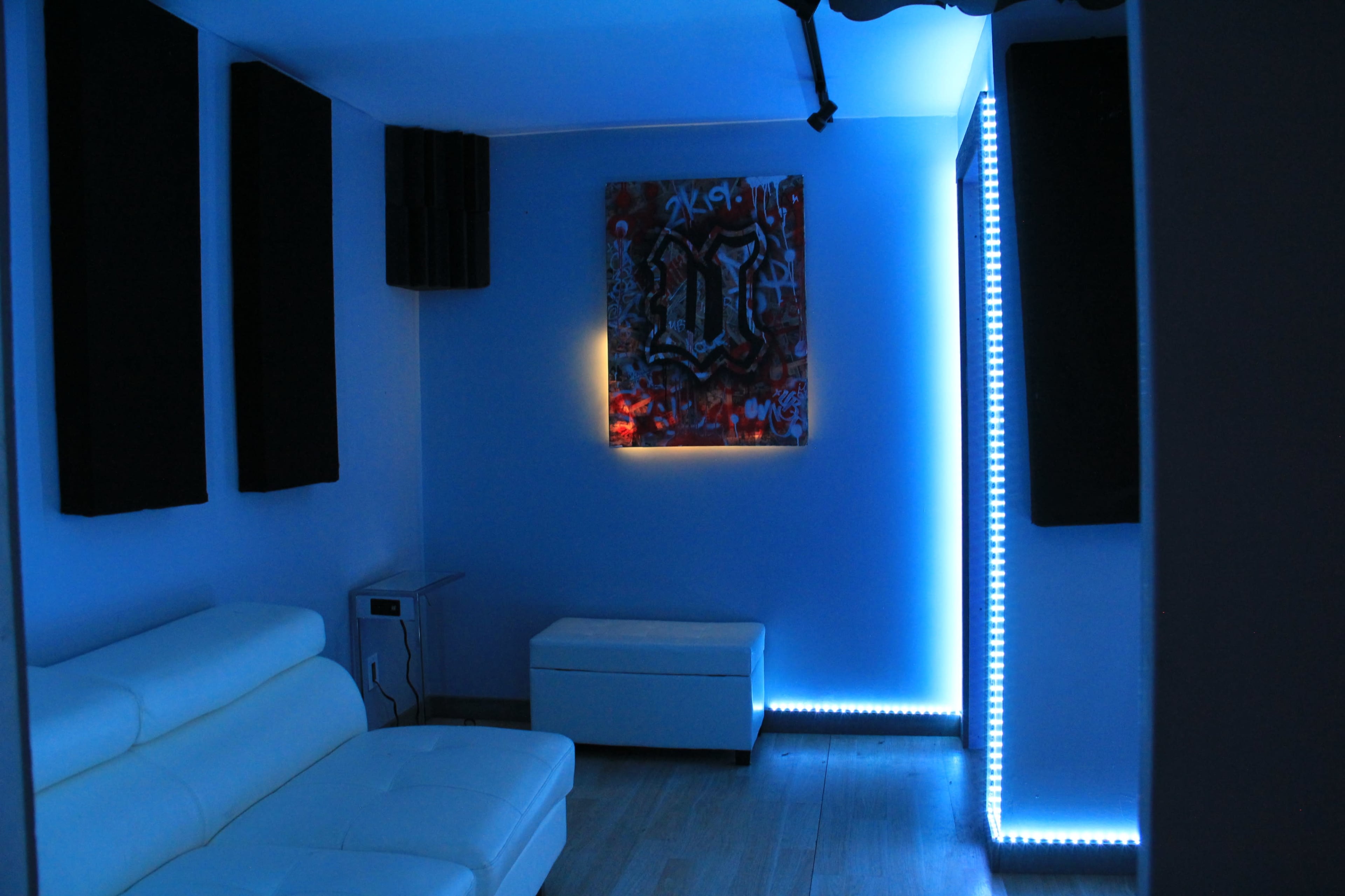 A room with blue lighting, featuring a white couch, a light-colored cabinet, and soundproofing panels on the walls.