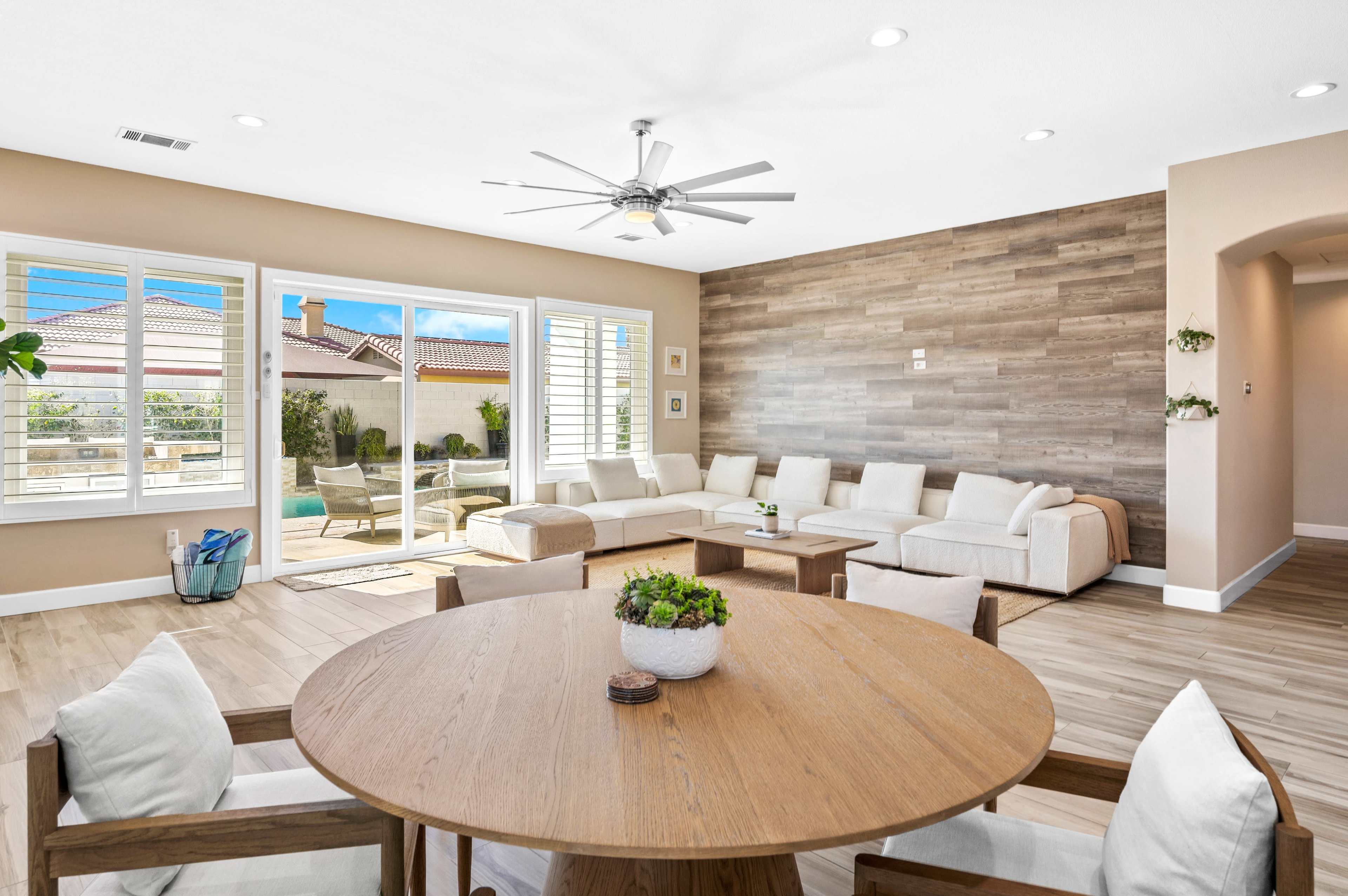 A modern living room features a circular dining table surrounded by chairs, with a sectional sofa and large windows overlooking an outdoor space.