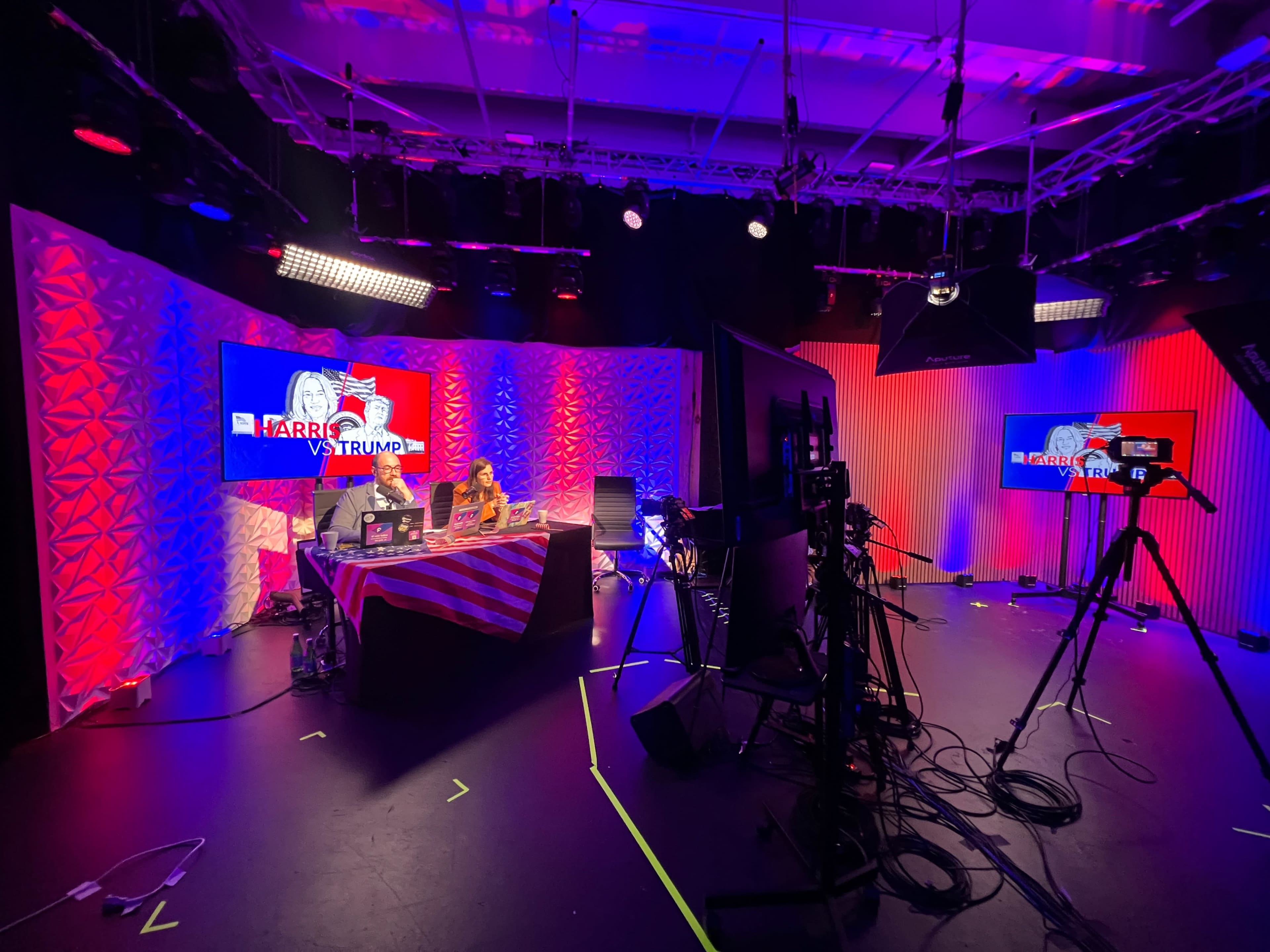 The image shows a television studio set with a brightly lit desk area, featuring screens displaying "Harris vs Trump," and cameras positioned for a broadcast.