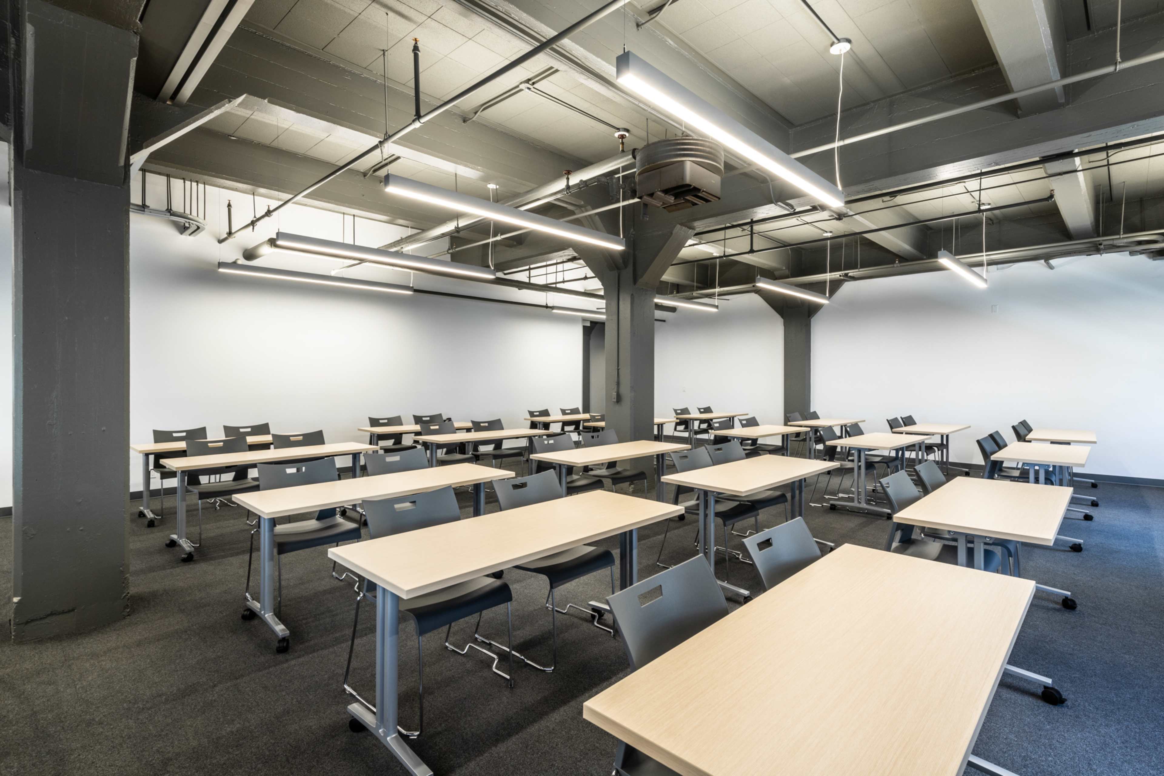 A spacious classroom featuring rows of tables and chairs arranged neatly, with gray walls and overhead lighting.