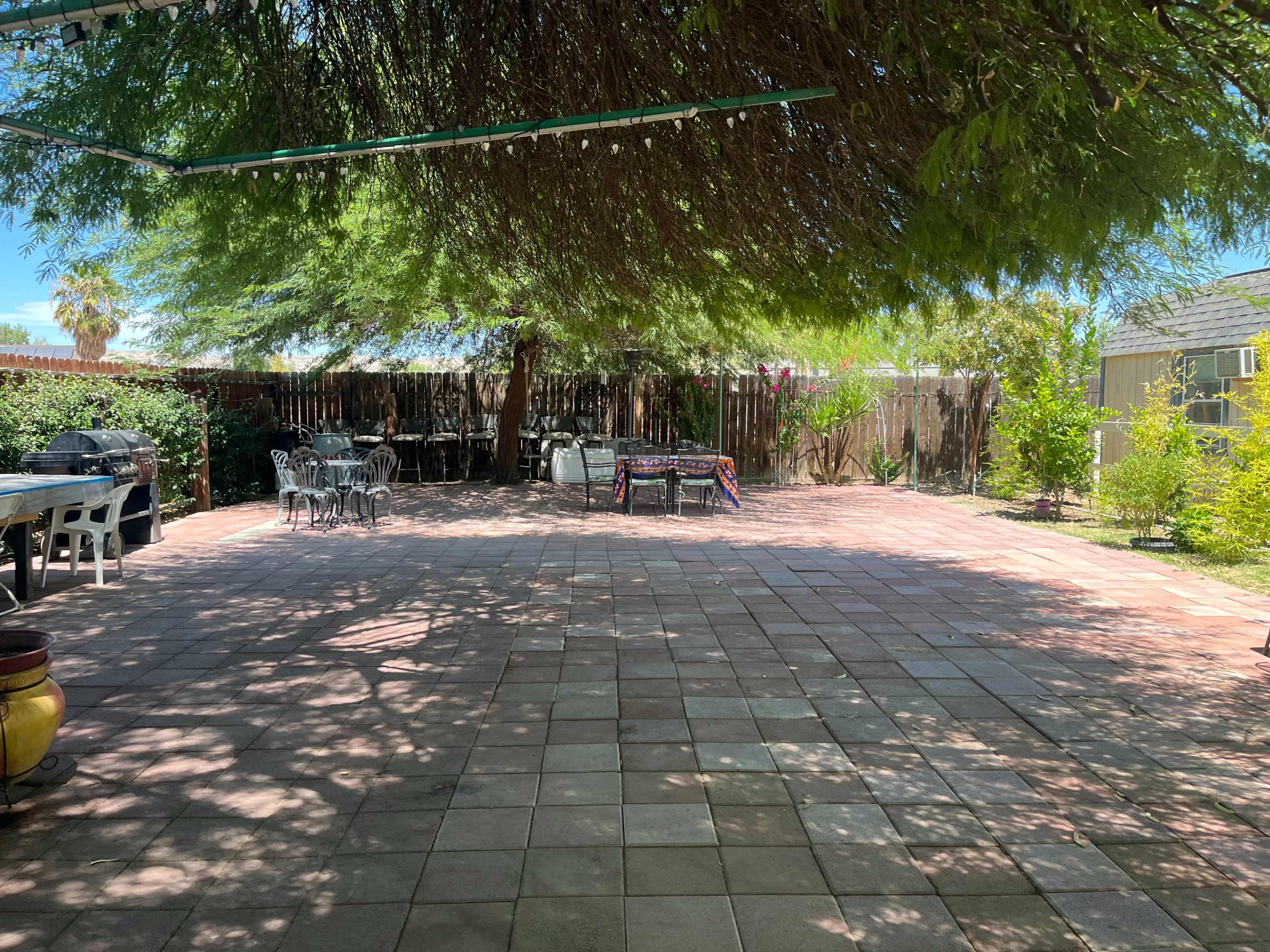 The image shows a spacious outdoor patio area with tiled flooring, surrounded by greenery and shaded by trees.