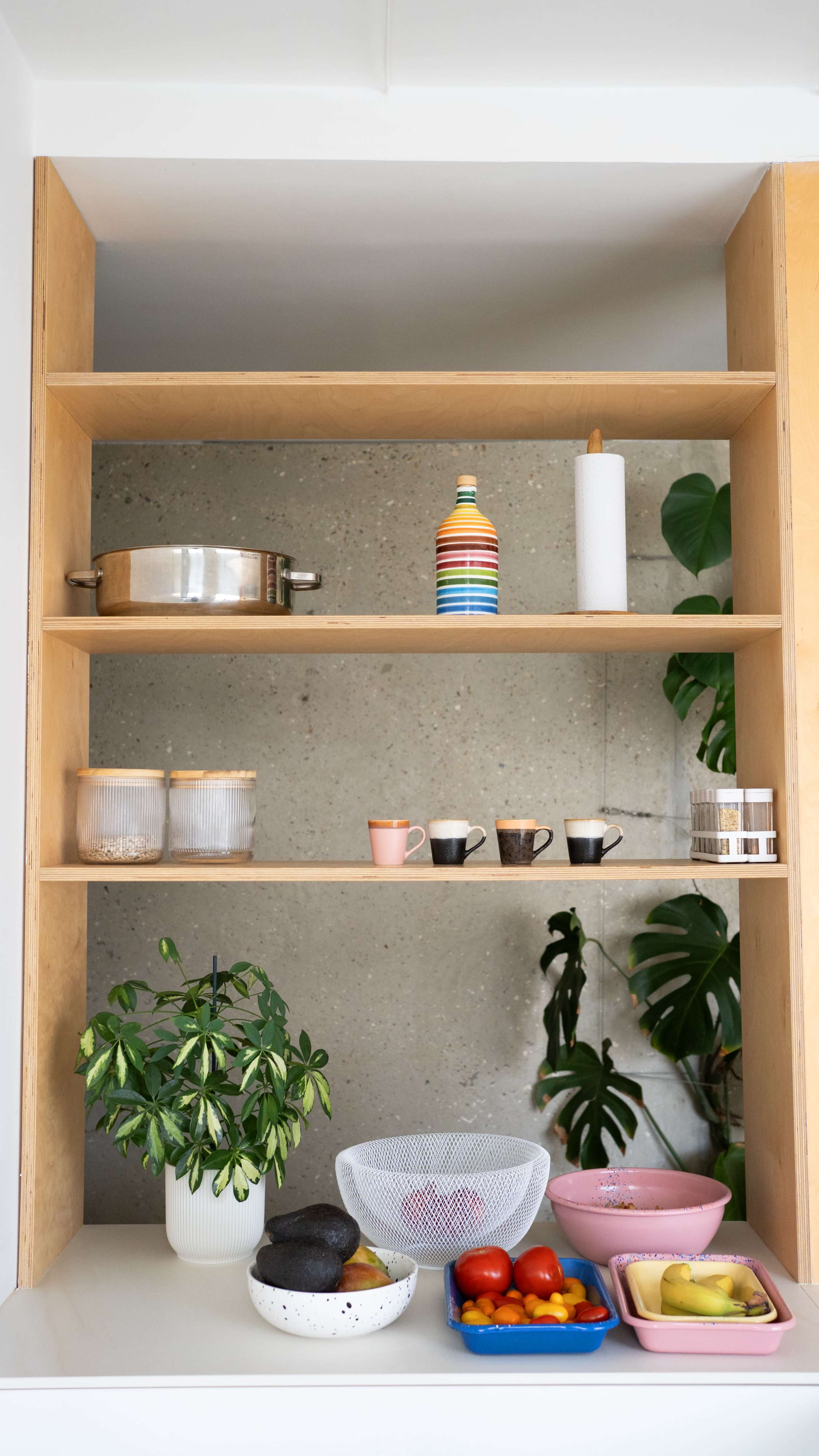 The image shows a wooden shelf with various kitchen items, including pots, cups, and bowls, alongside a green plant and colorful fruits arranged in bowls.