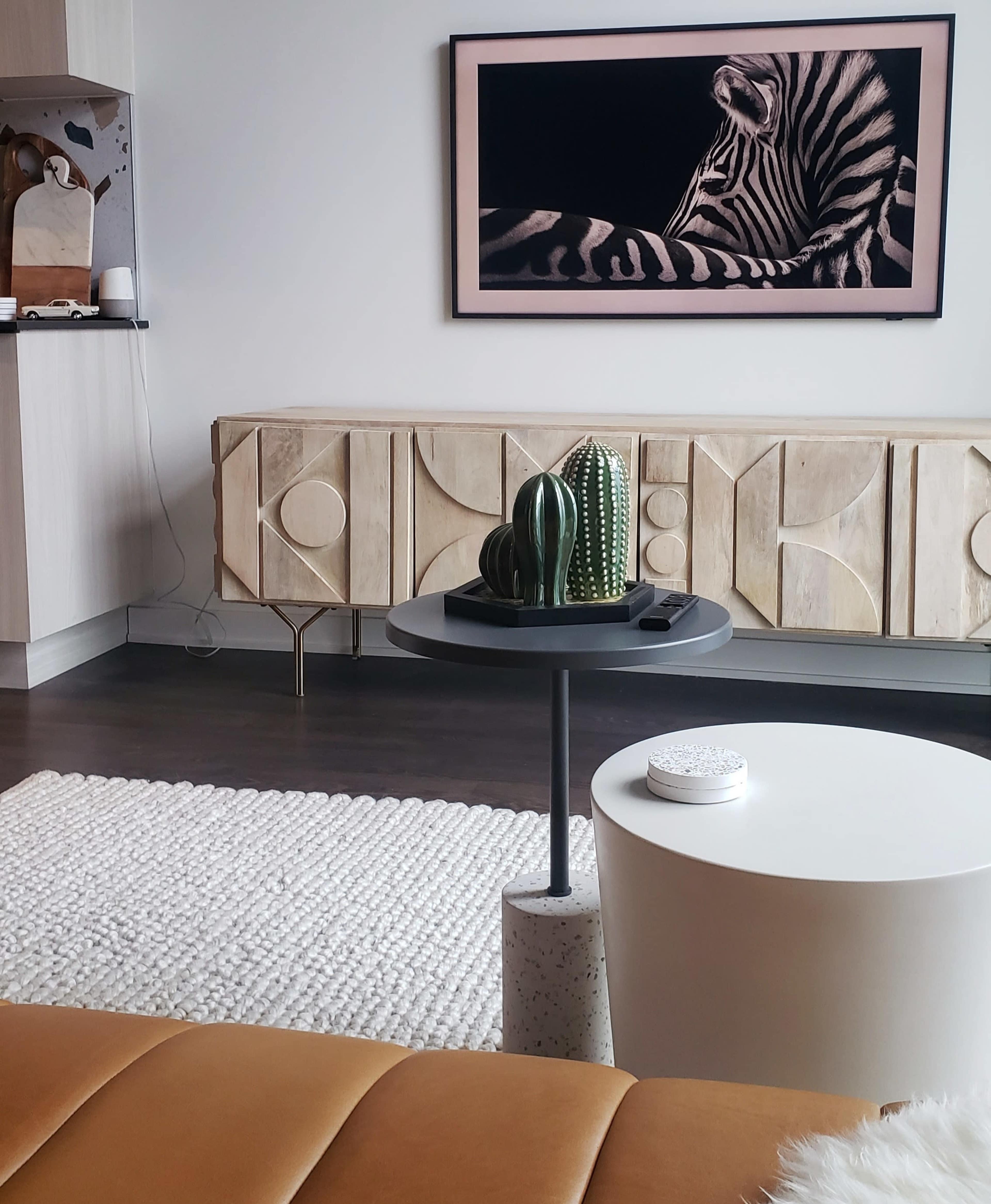 A modern living room with a brown sofa, a circular side table holding decorative items, and a large zebra-print artwork displayed on the wall above a wooden cabinet.
