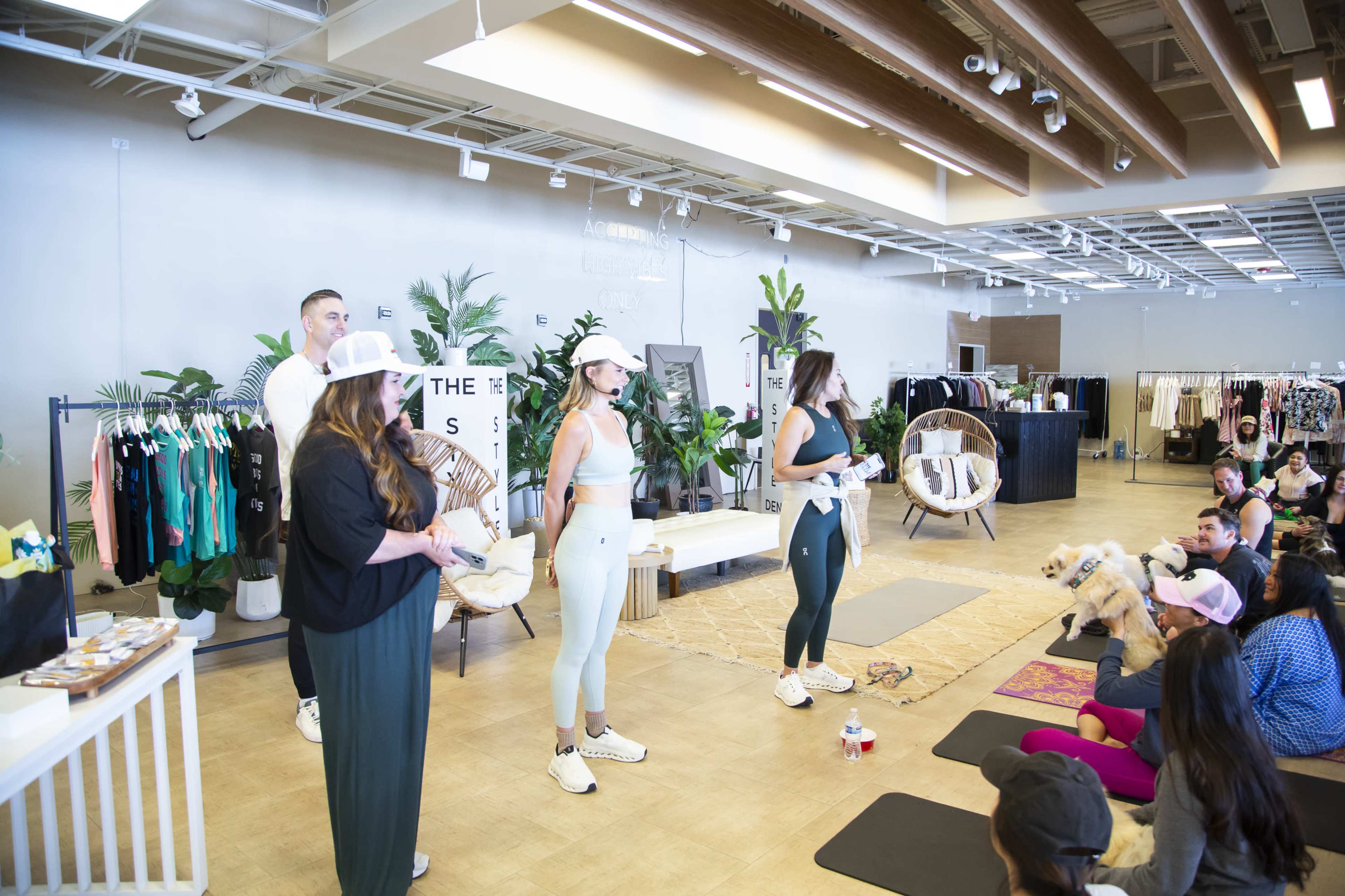 A fitness event is taking place in a modern store with four presenters demonstrating exercises to an audience sitting on yoga mats.