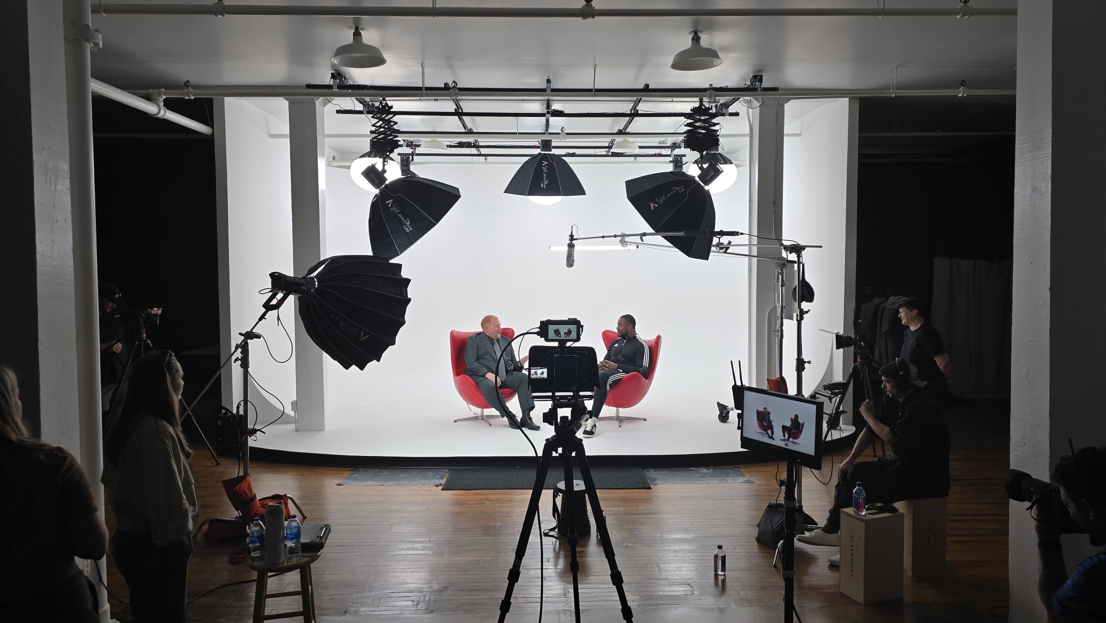 Two people sit in red chairs against a white backdrop in a well-lit studio, surrounded by camera equipment and lighting setups.