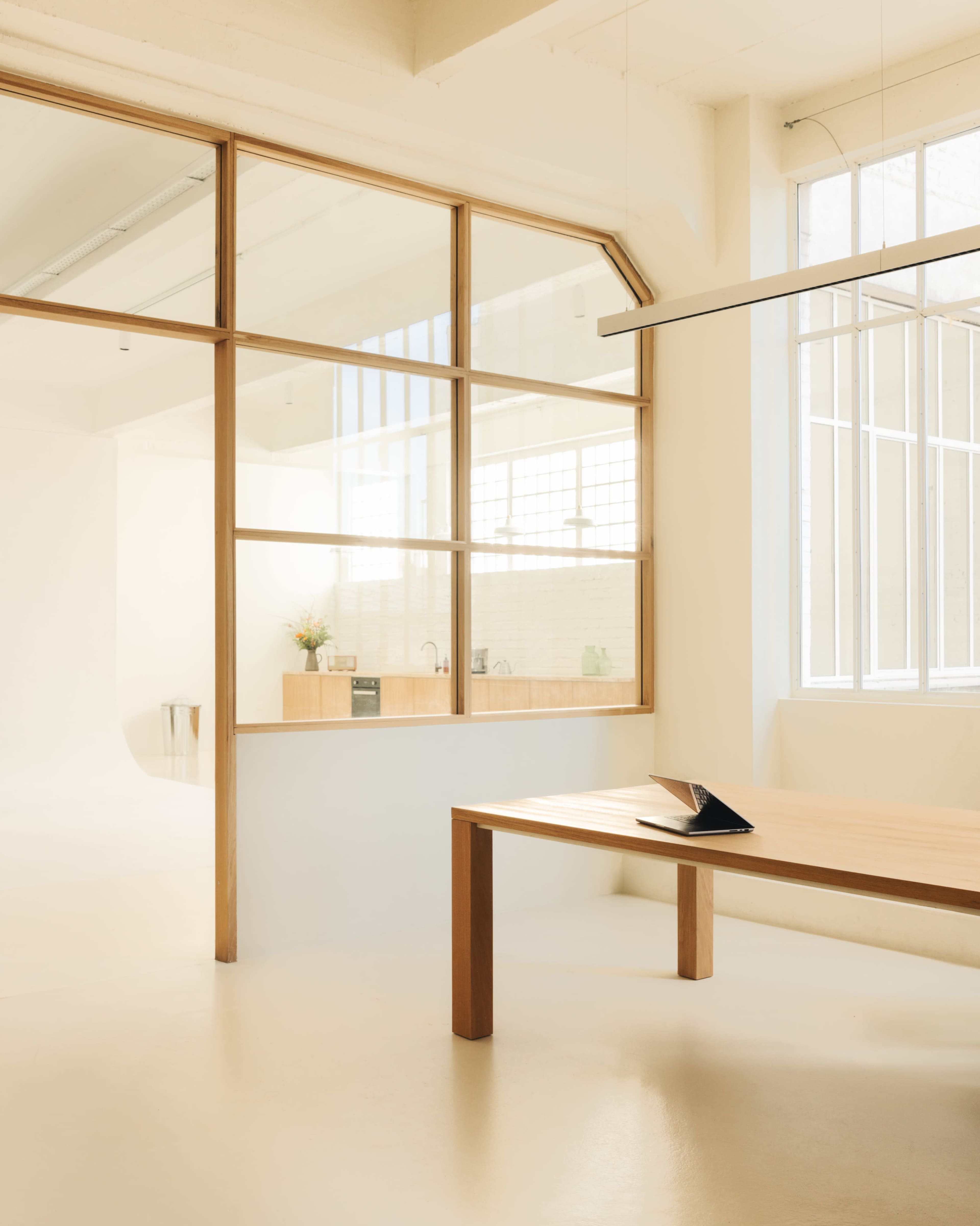 A minimalist interior features a large wooden table in the foreground and a glass partition separating a bright kitchen area in the background.