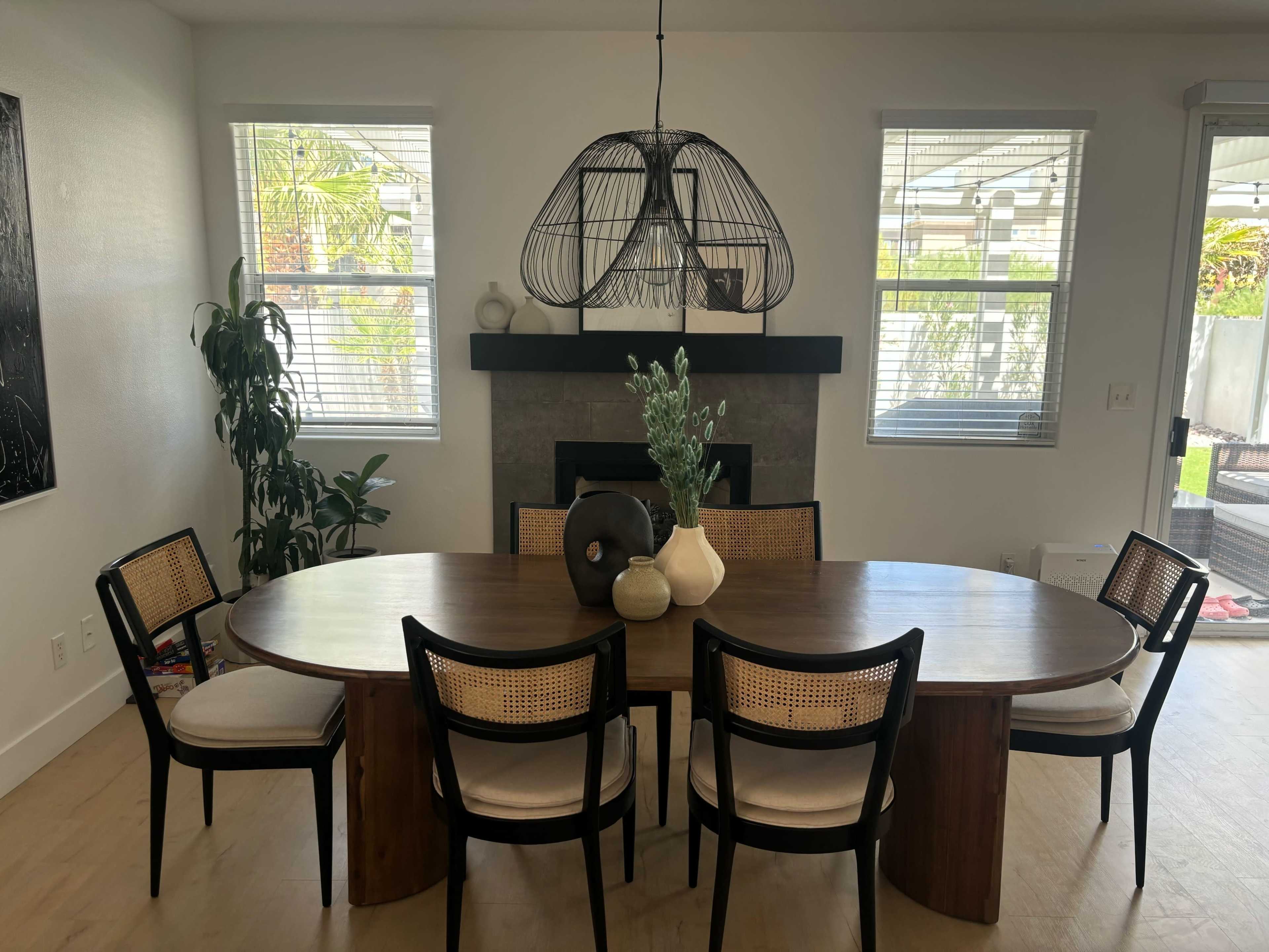 A round wooden dining table with six chairs is positioned in a bright room featuring a large pendant light and a fireplace.