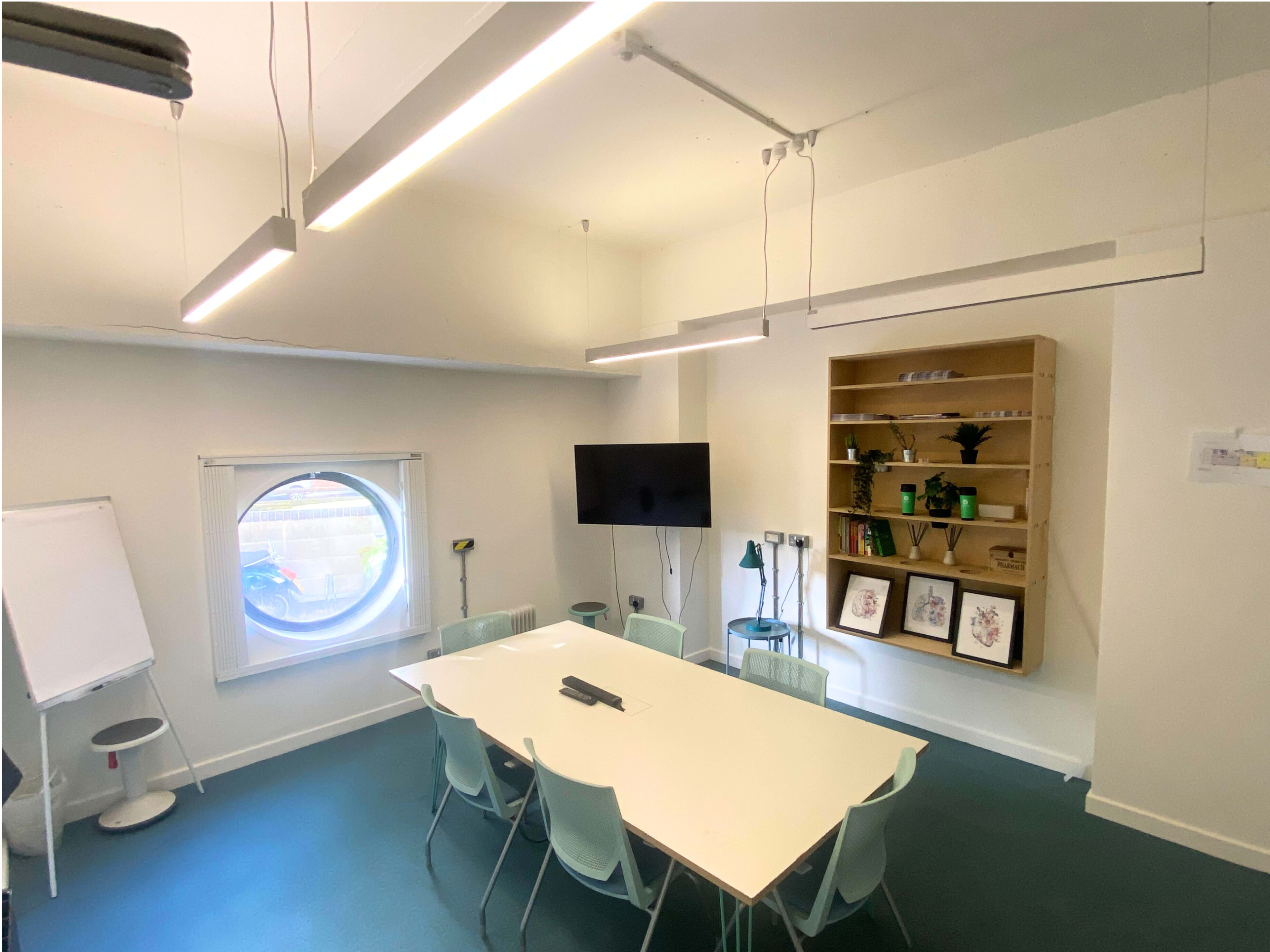 The image shows a modern meeting room featuring a large white table surrounded by chairs, a circular window, a television on the wall, and shelves with decorative items.