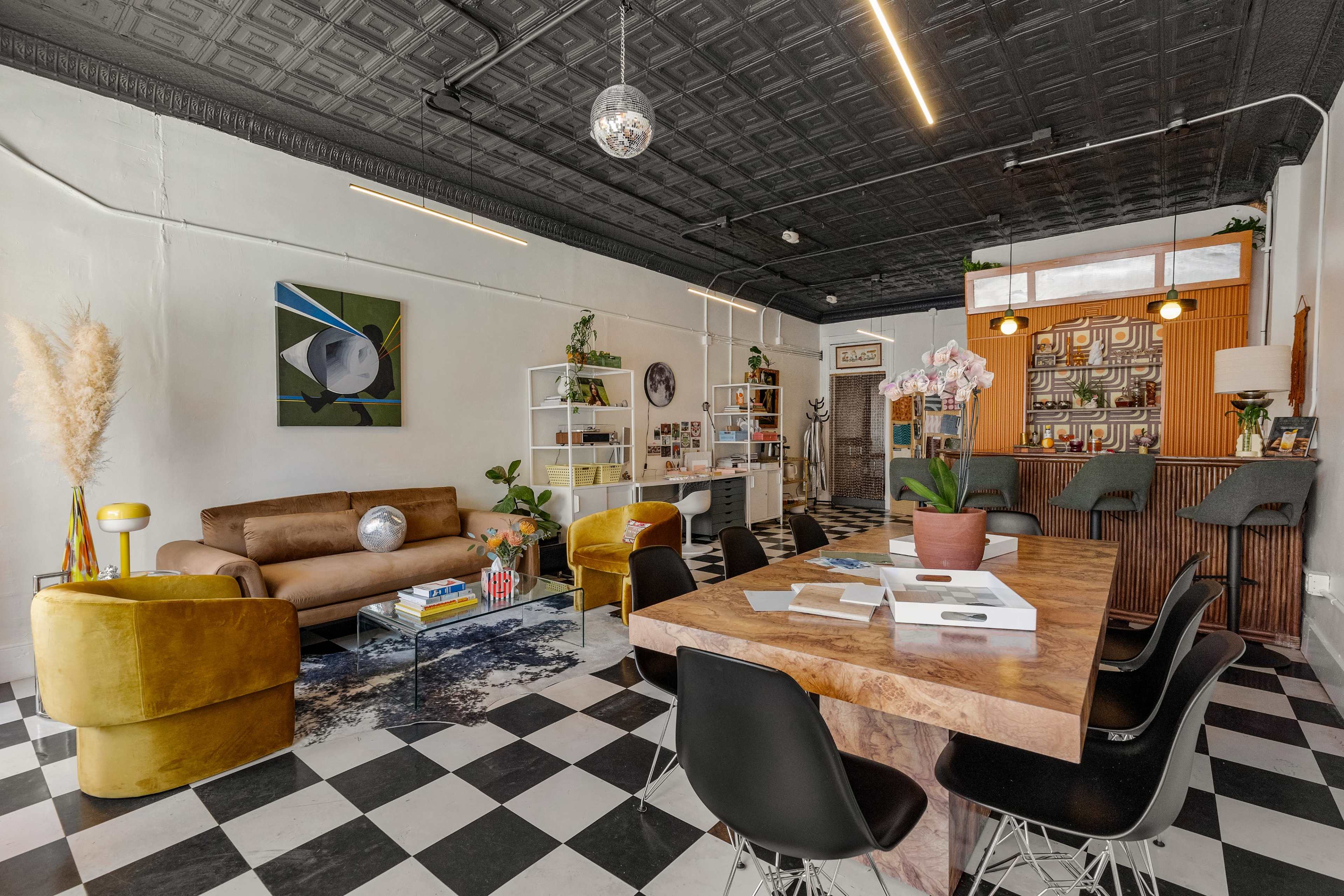 The image shows a modern, eclectic interior space with a brown sofa, a large wooden table surrounded by black chairs, and various decorative plants and artworks.