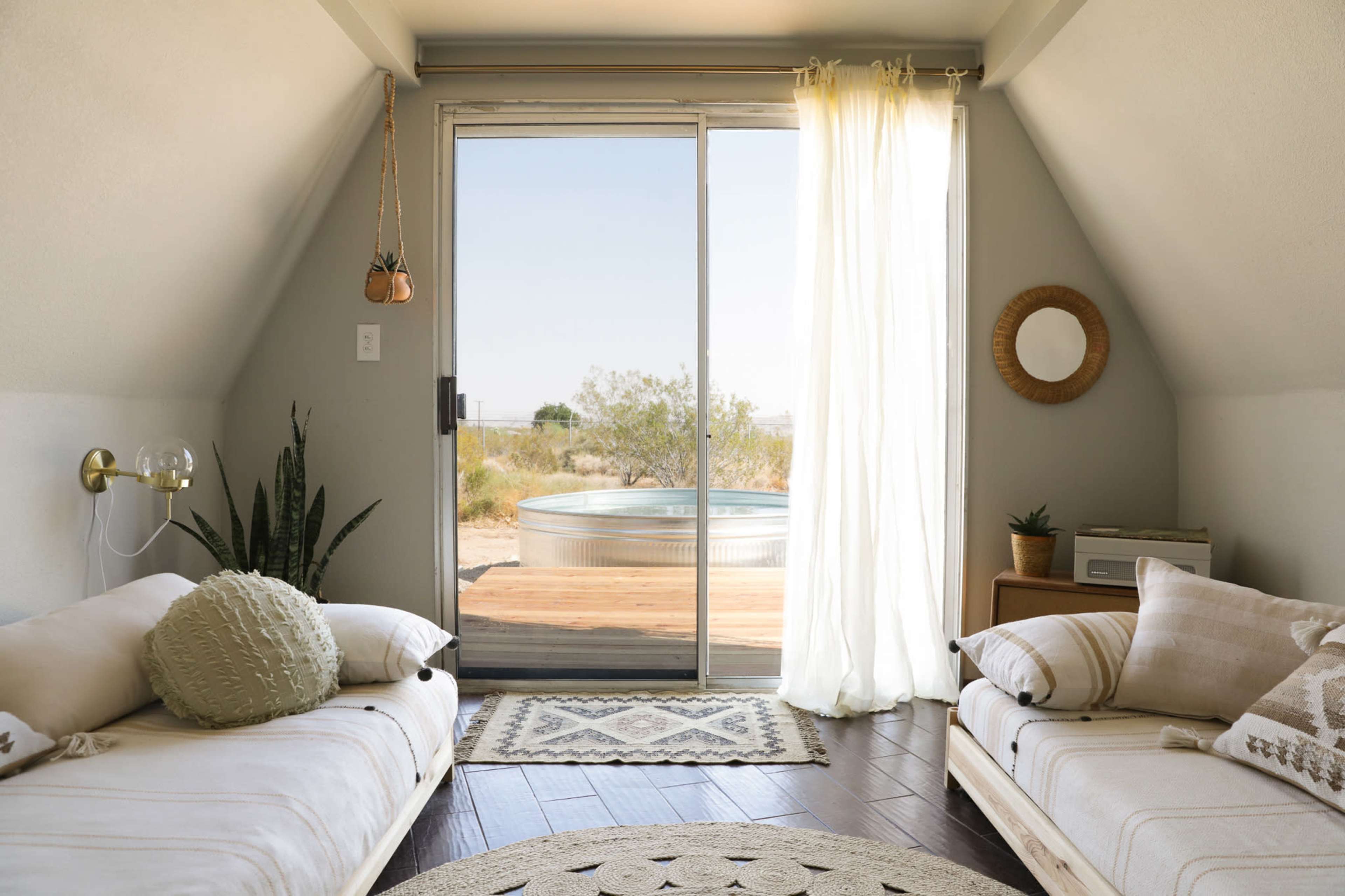 A cozy, minimalist room features two beds, a sliding glass door leading to an outdoor area, and natural light flowing in.