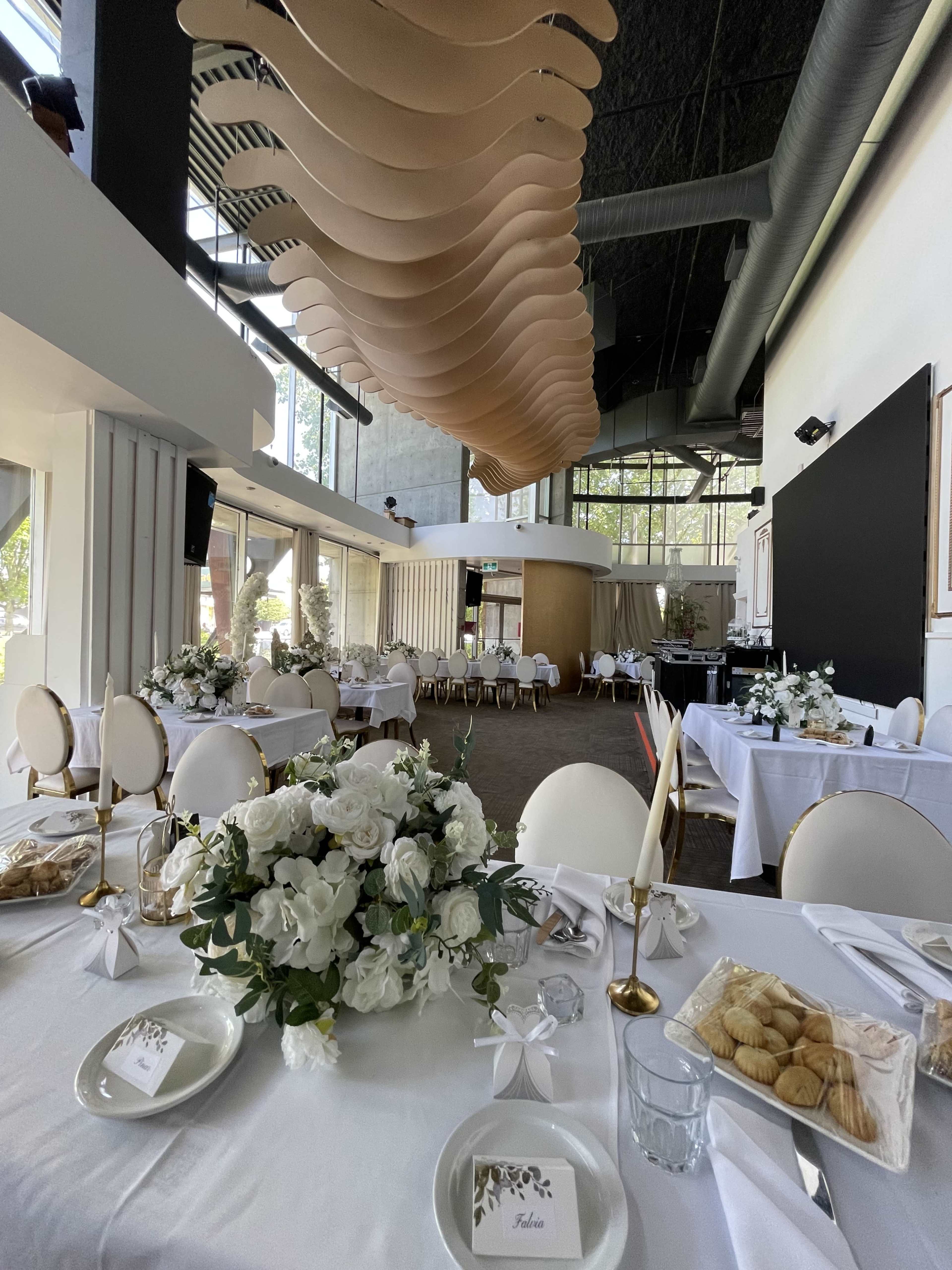 The image shows a banquet hall with elegantly set tables featuring floral centerpieces and a modern ceiling design.