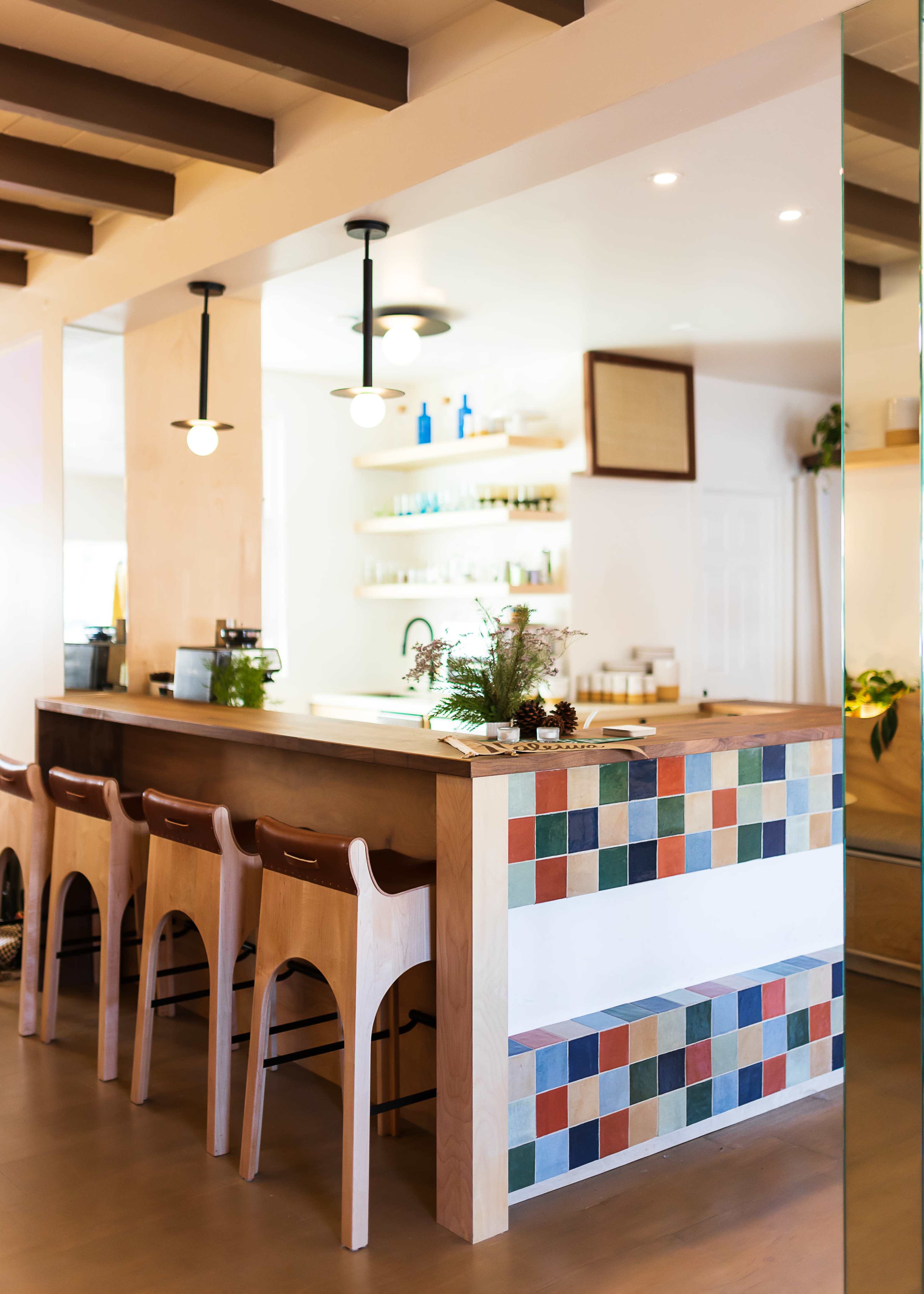A clean, modern café interior features a light wooden bar with colorful tiled accents and a row of wooden stools.