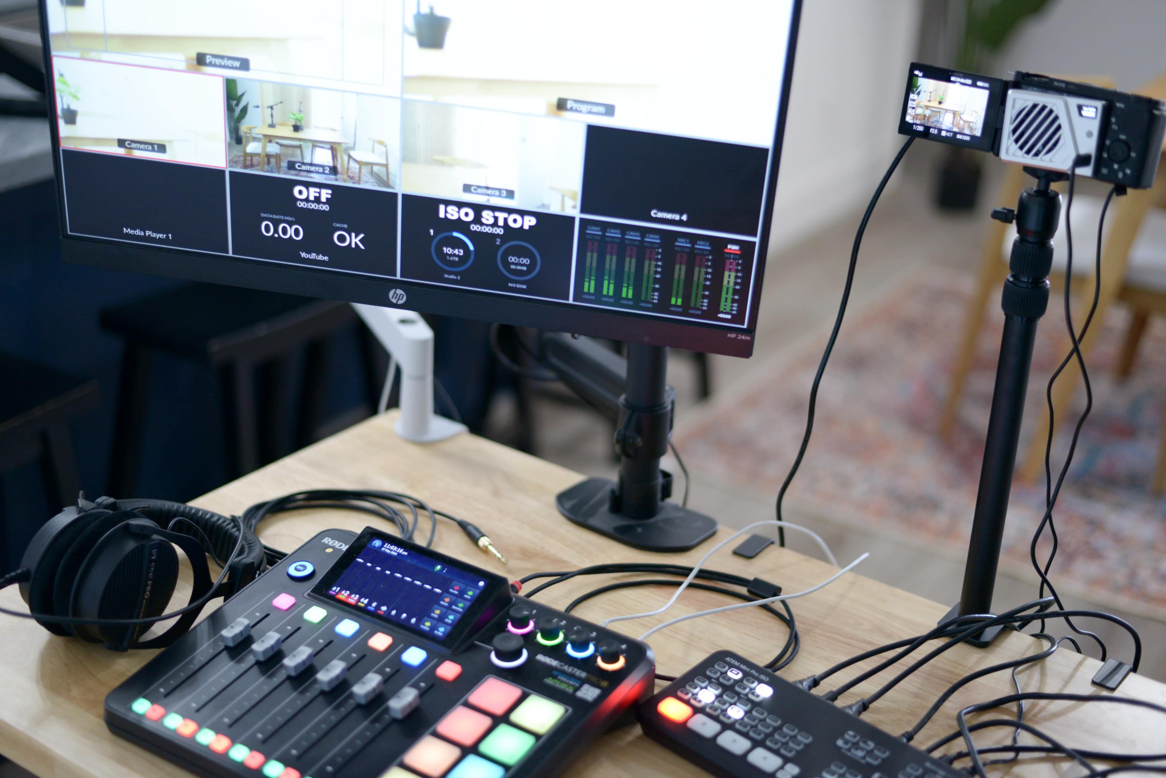 A computer monitor displays multiple camera angles while a audio mixer, a microphone, and headphones are arranged on a wooden desk.